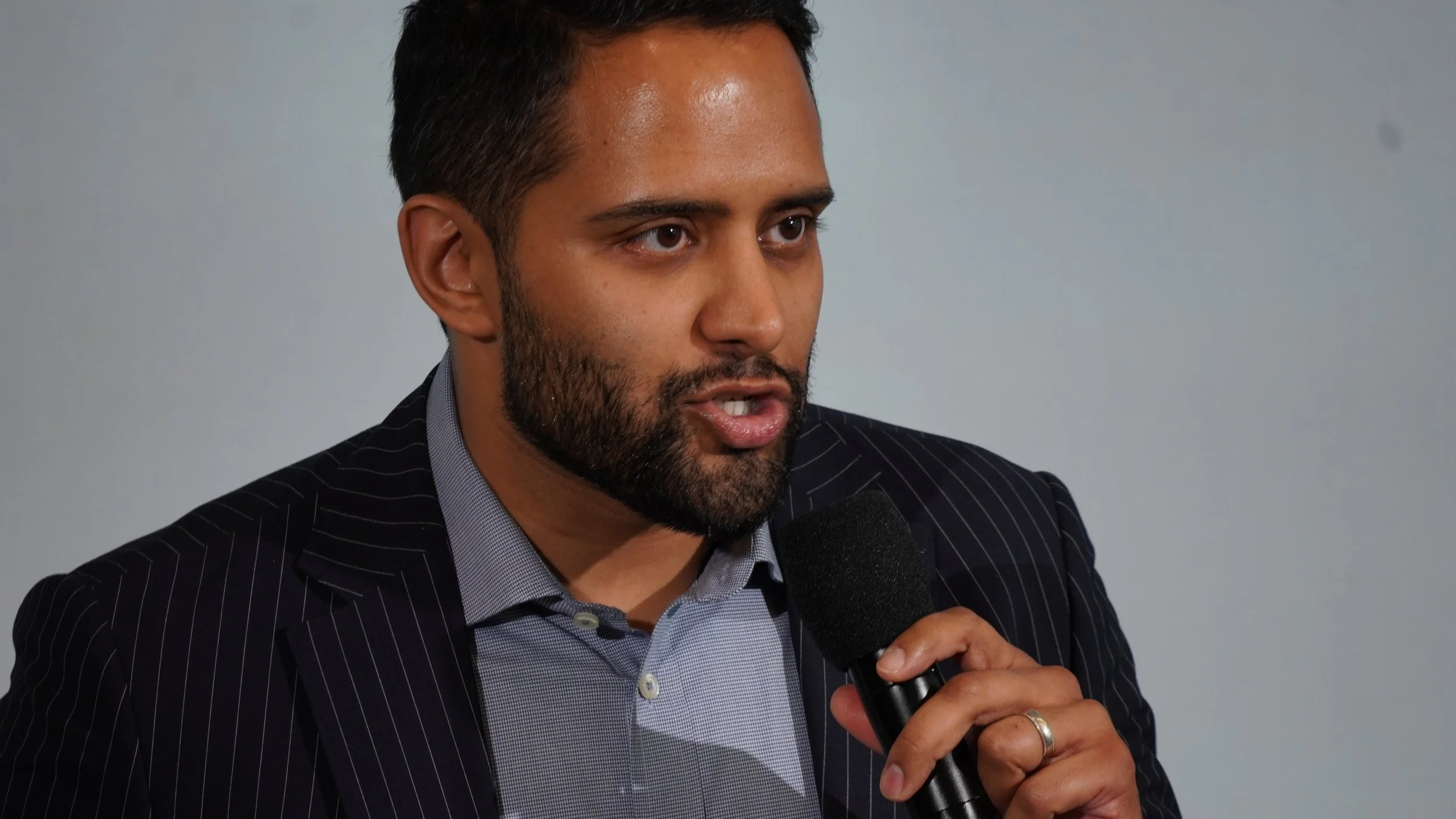 A man with dark hair and a beard, wearing a dark pinstripe suit and a light-colored shirt, speaking into a black microphone.