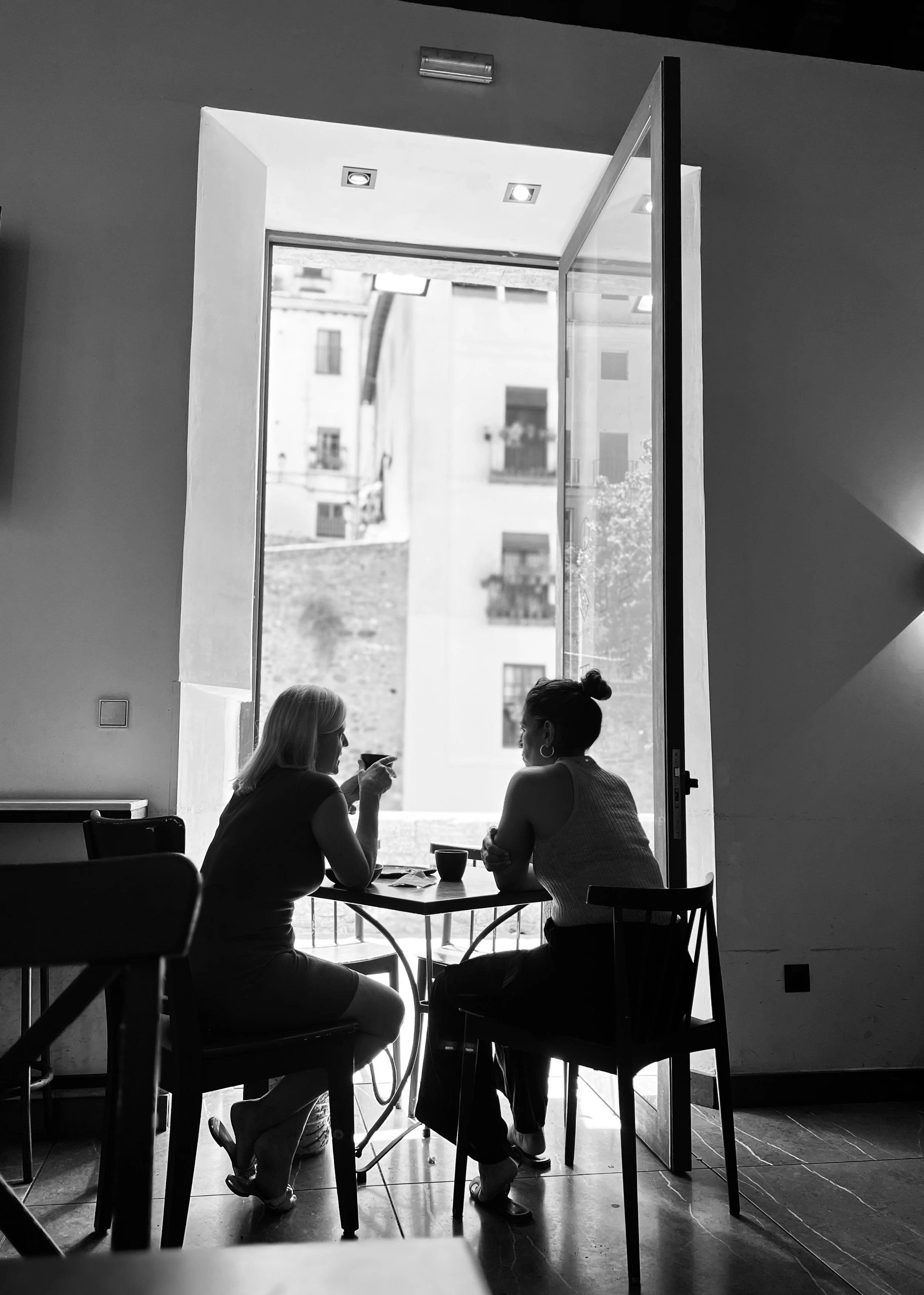 Two women sitting at a small table near a large open window in a cafe, engaged in conversation. It is a black and white photo.