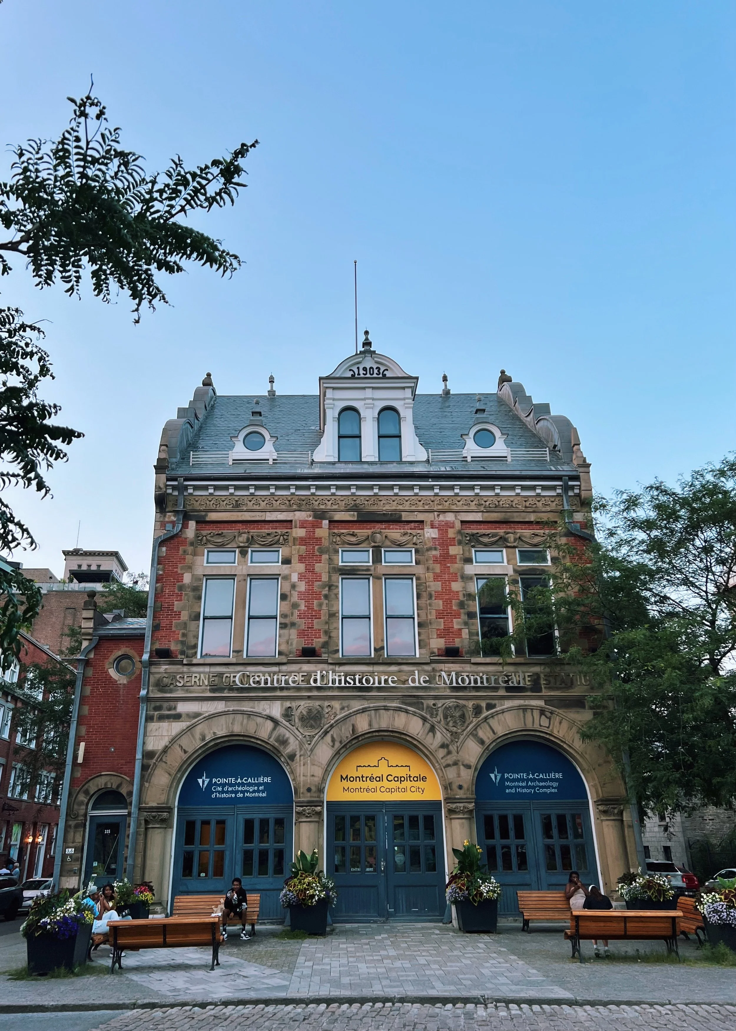 Historic building with arched windows and a sign that reads 'Centre d'histoire de Montréal' and 'Montreal Capitale City.' Exterior featuring decorative stonework, two blue doors, benches and potted plants in front, with a few people sitting on benche