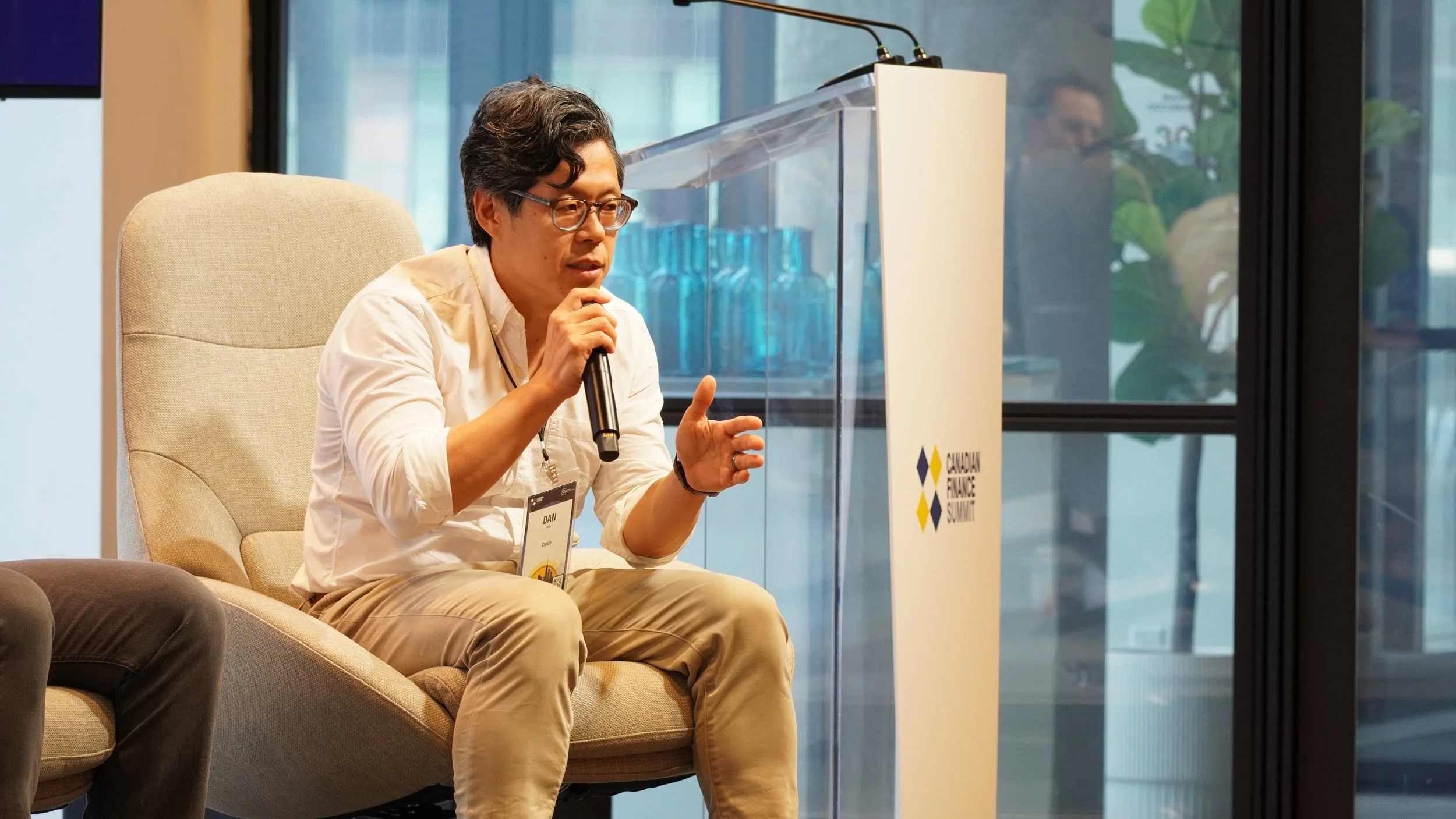 A man with curly dark hair, glasses, wearing a white shirt and tan pants, is sitting on a beige chair, speaking into a microphone, at the Canadian Finance Summit. There is a transparent podium with the summit logo beside him and a large window in the