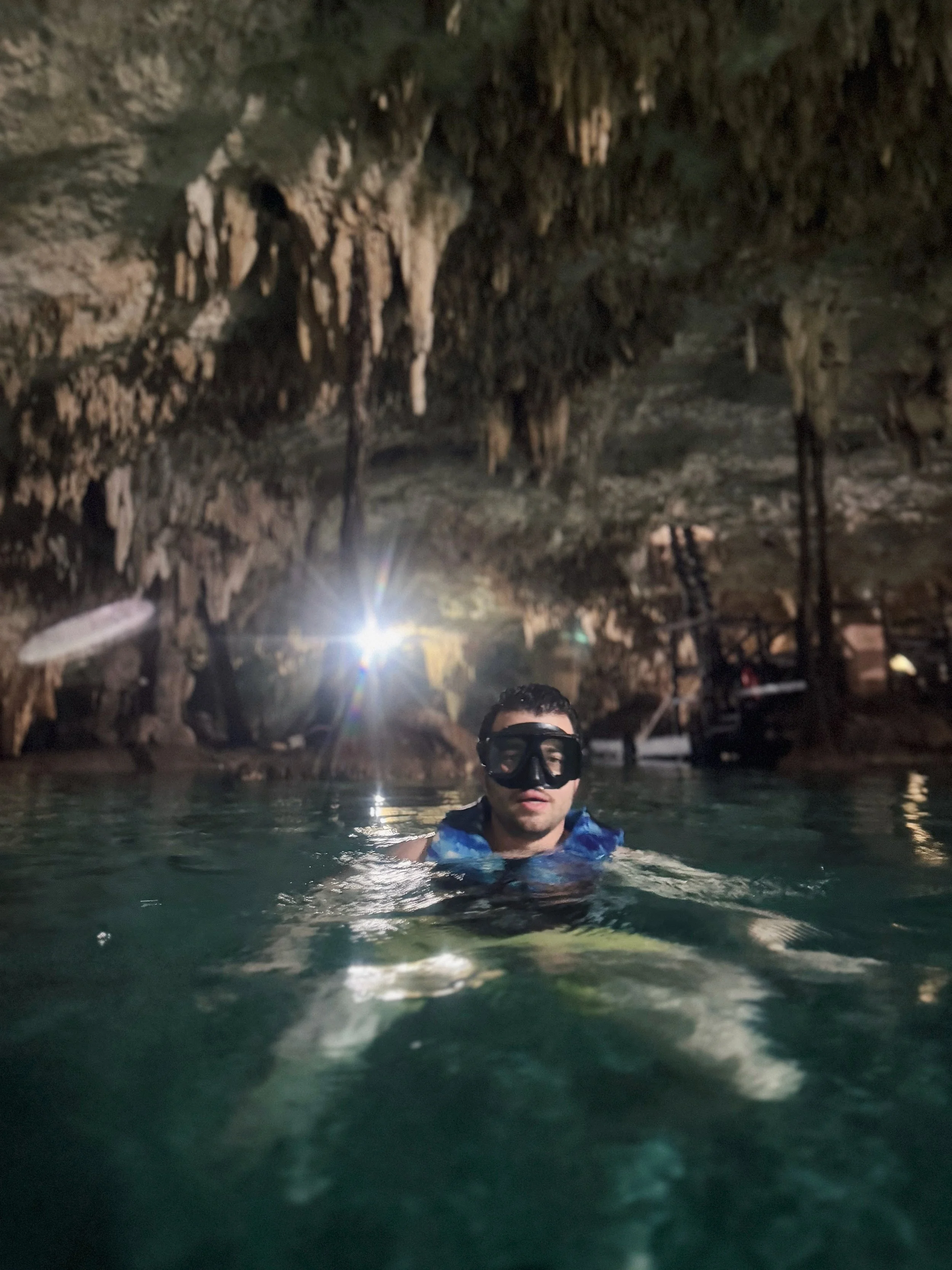 A man wearing a wetsuit and snorkeling mask swimming in a cave with stalactites and stalagmites, with a light shining from behind.