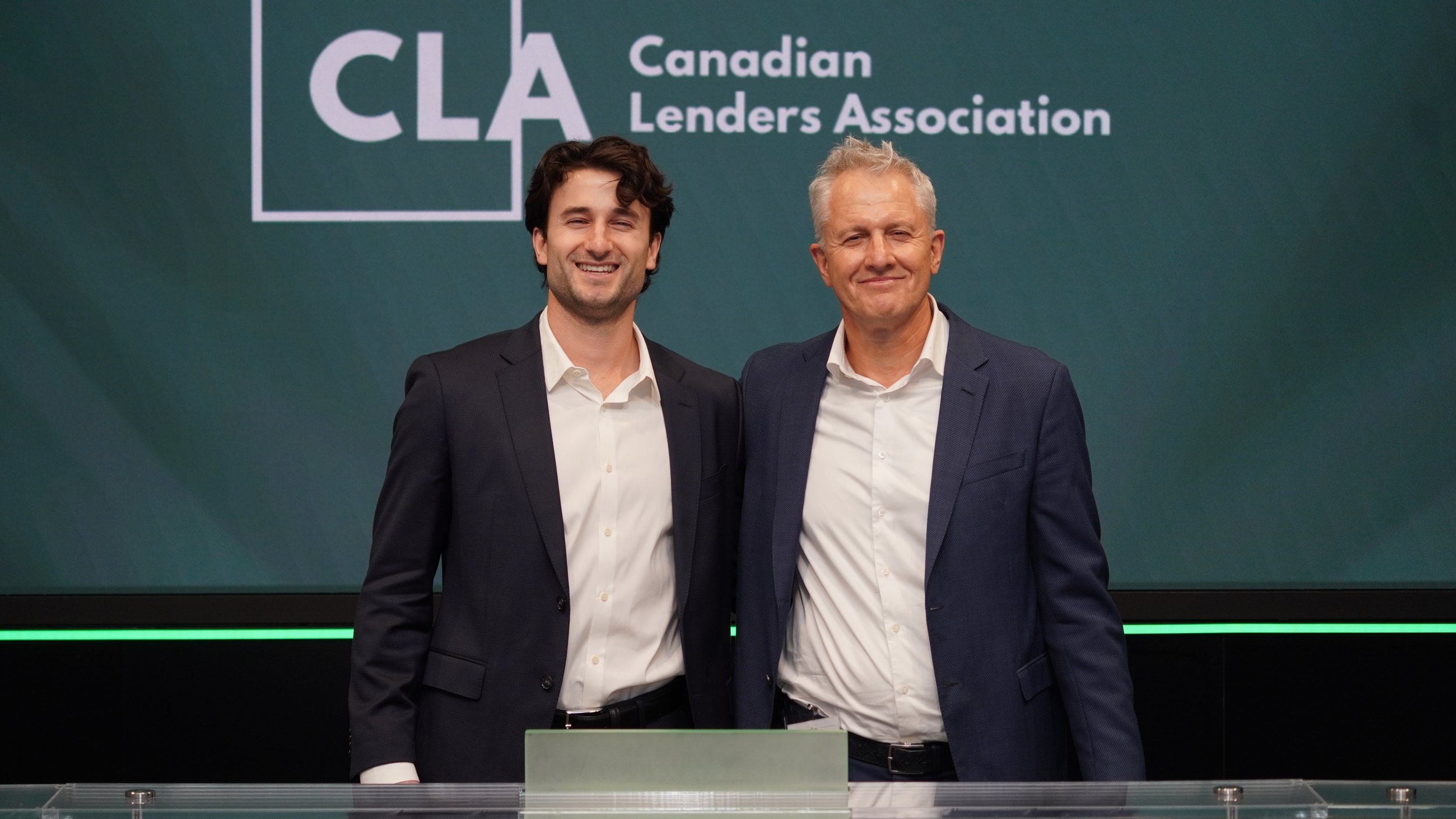 Two men in business suits standing side by side at a conference, smiling, with a green display behind them that reads 'Canadian Lenders Association' and the abbreviation 'CLA'.