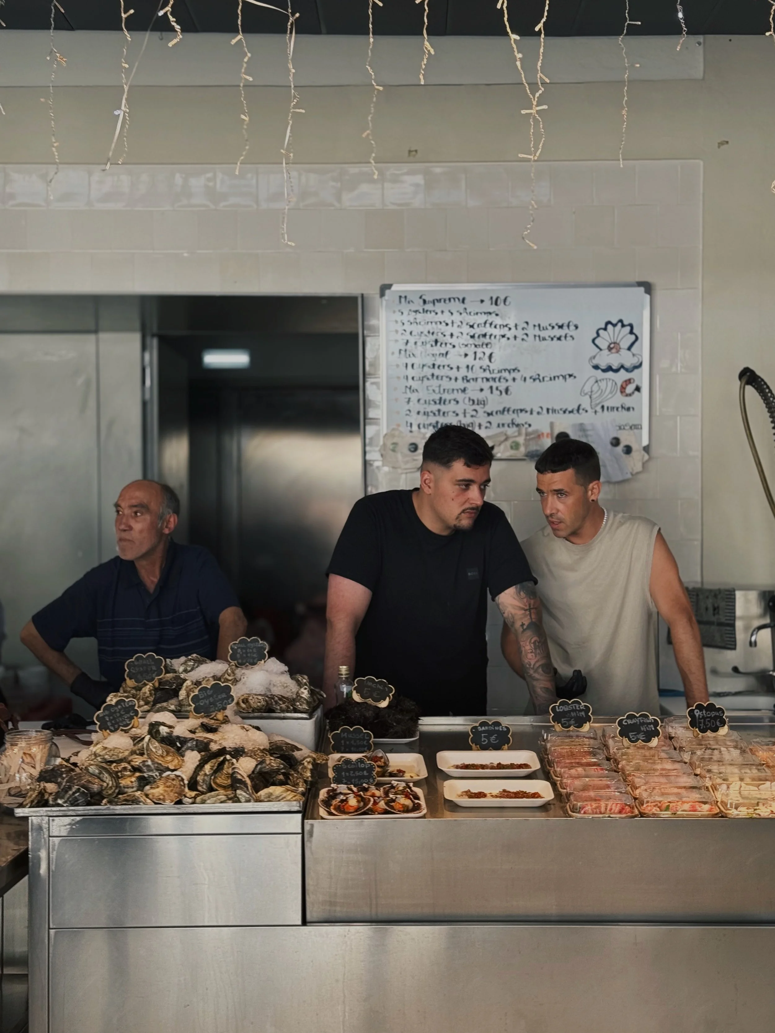 Three men behind a seafood counter with oysters and other seafood on display, amid a fish market or seafood shop setting.