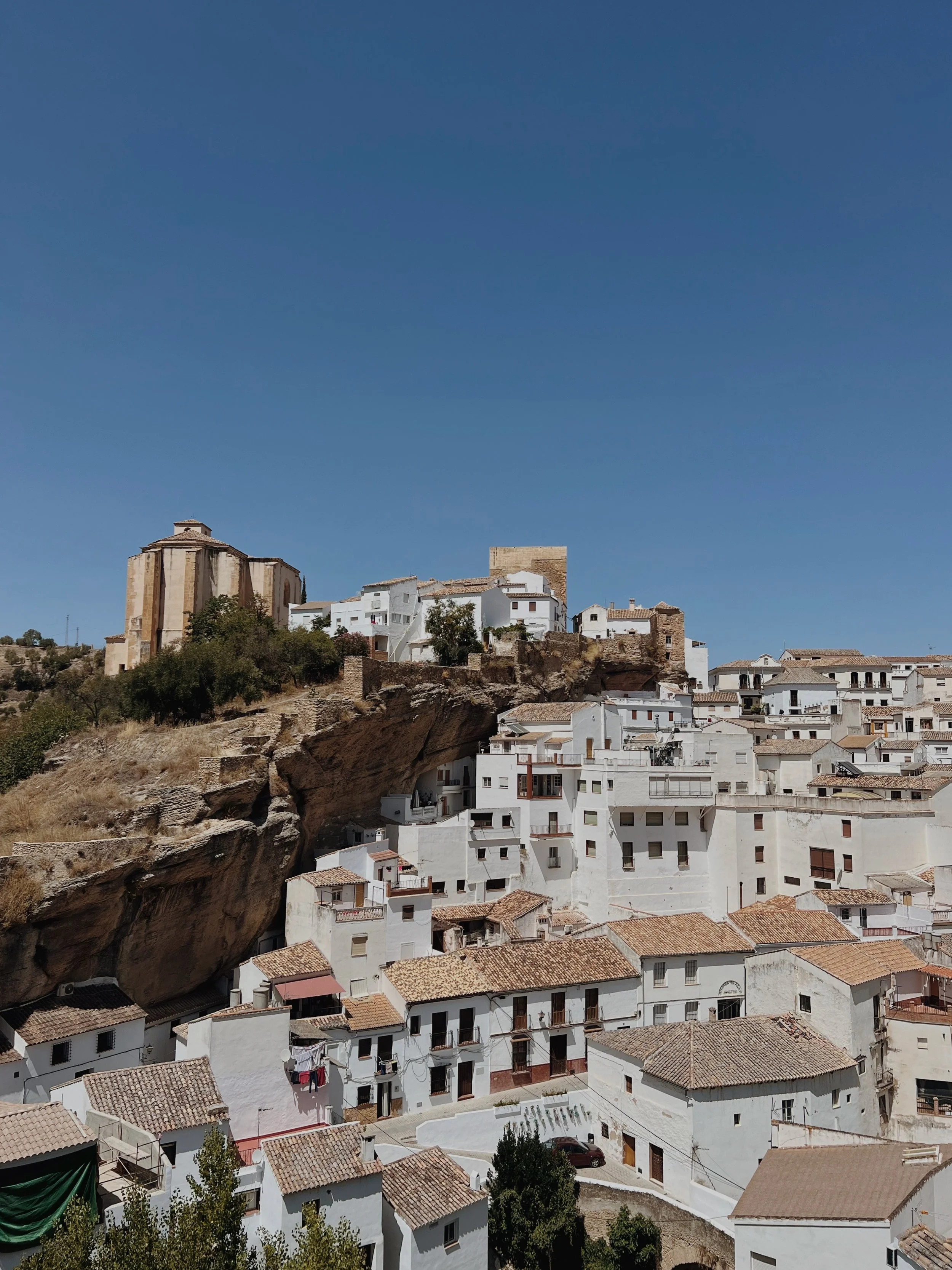 A hillside town with white buildings and terracotta rooftops built on a rocky slope, under a clear blue sky.