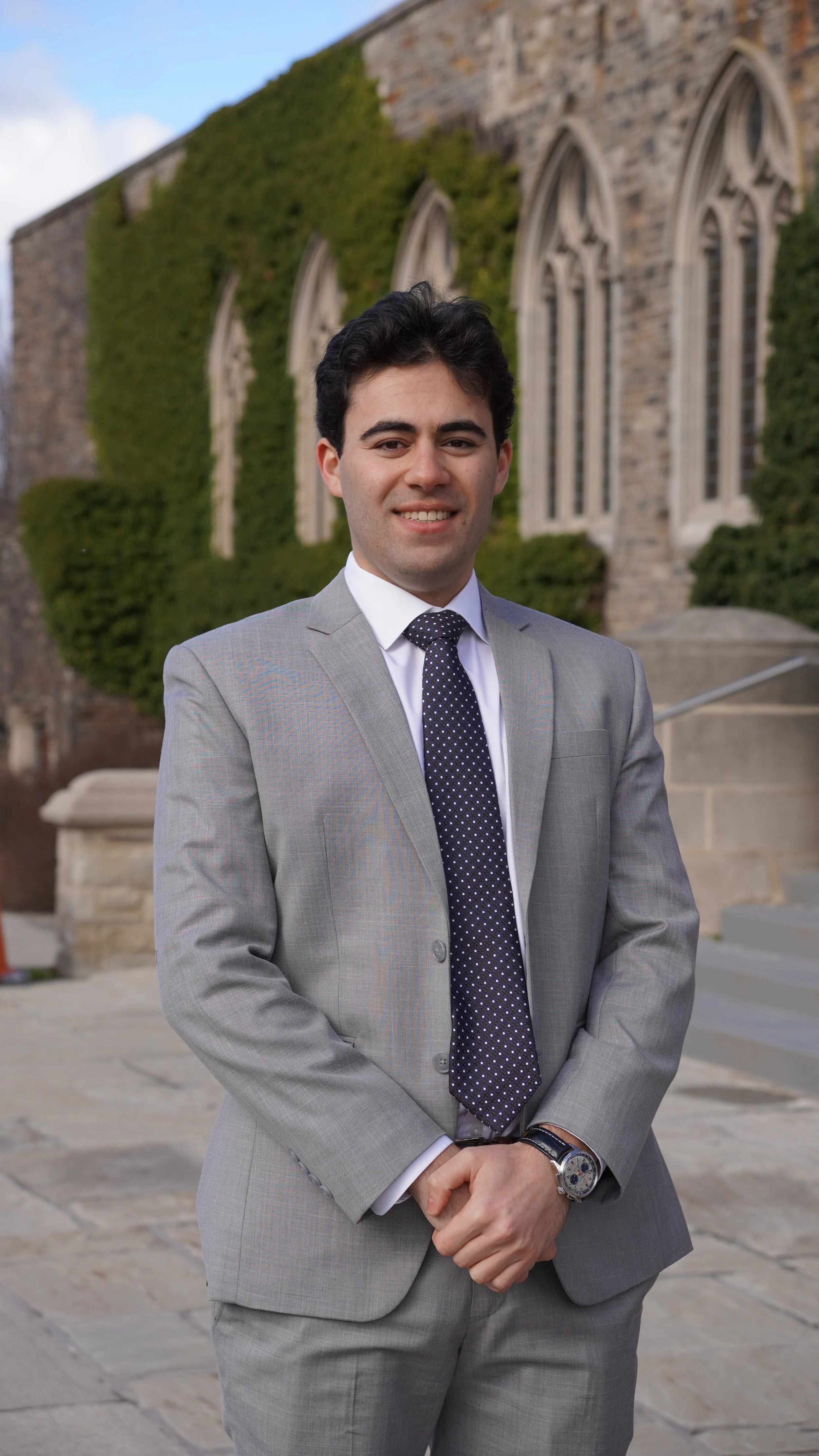 A young man in a gray suit with a dark polka dot tie standing in front of a historic stone building with arched windows and ivy growth.
