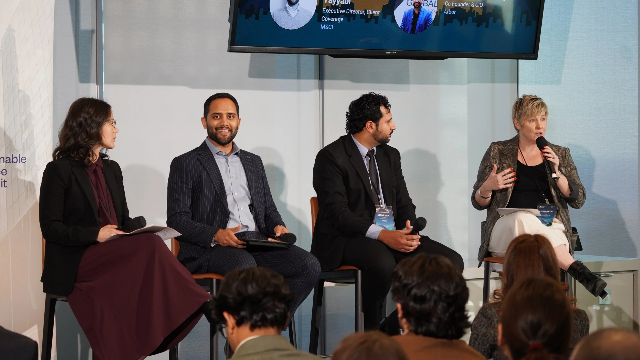 A panel of four professionals, three men and one woman, seated on stage during a discussion at a conference or seminar, with an audience watching.