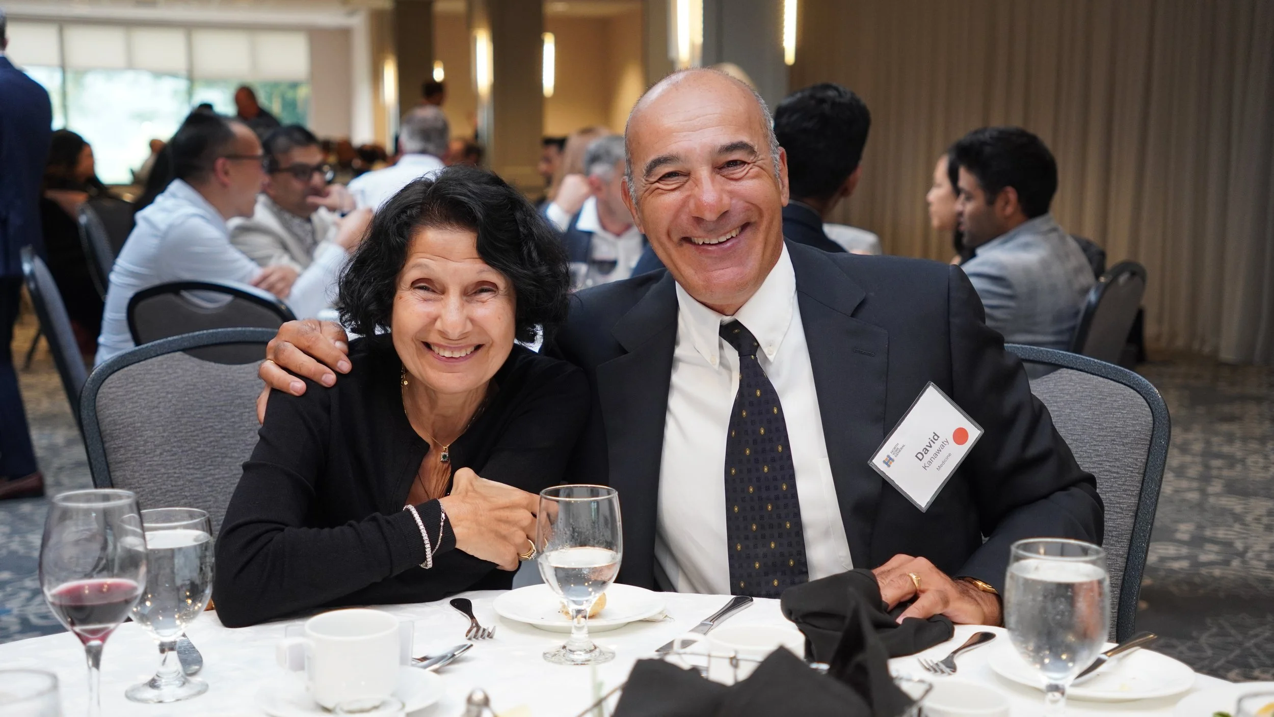Two smiling people, a woman and a man, sitting at a banquet table with glasses, plates, and silverware, in a large, well-lit event hall. The woman has dark curly hair and the man is bald with a name badge that reads 'David.'