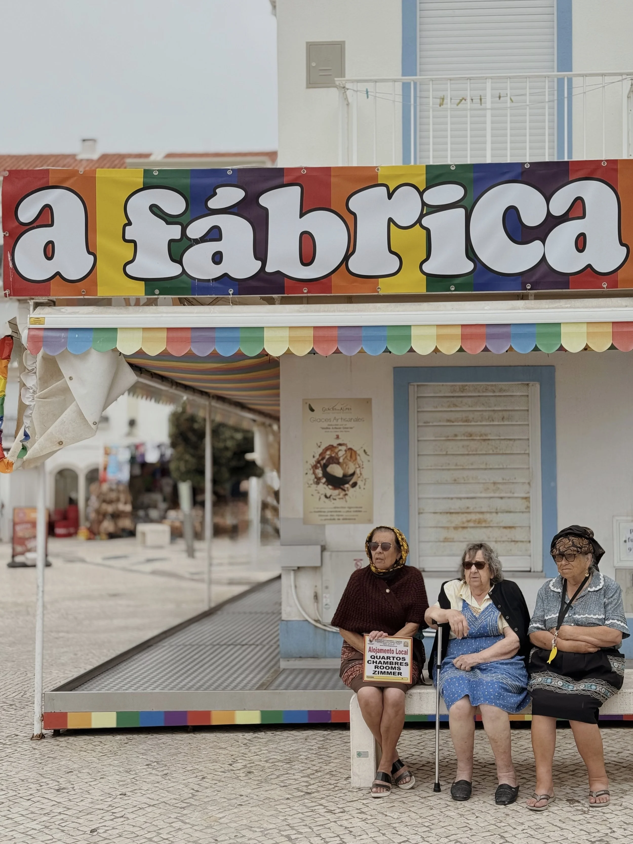 Three elderly women sitting on a bench in front of a colorful shop with a rainbow sign that reads 'a fábrica'. The women are dressed in dresses and sunglasses, and one has a cane, while the shop appears to sell local food and accommodations.