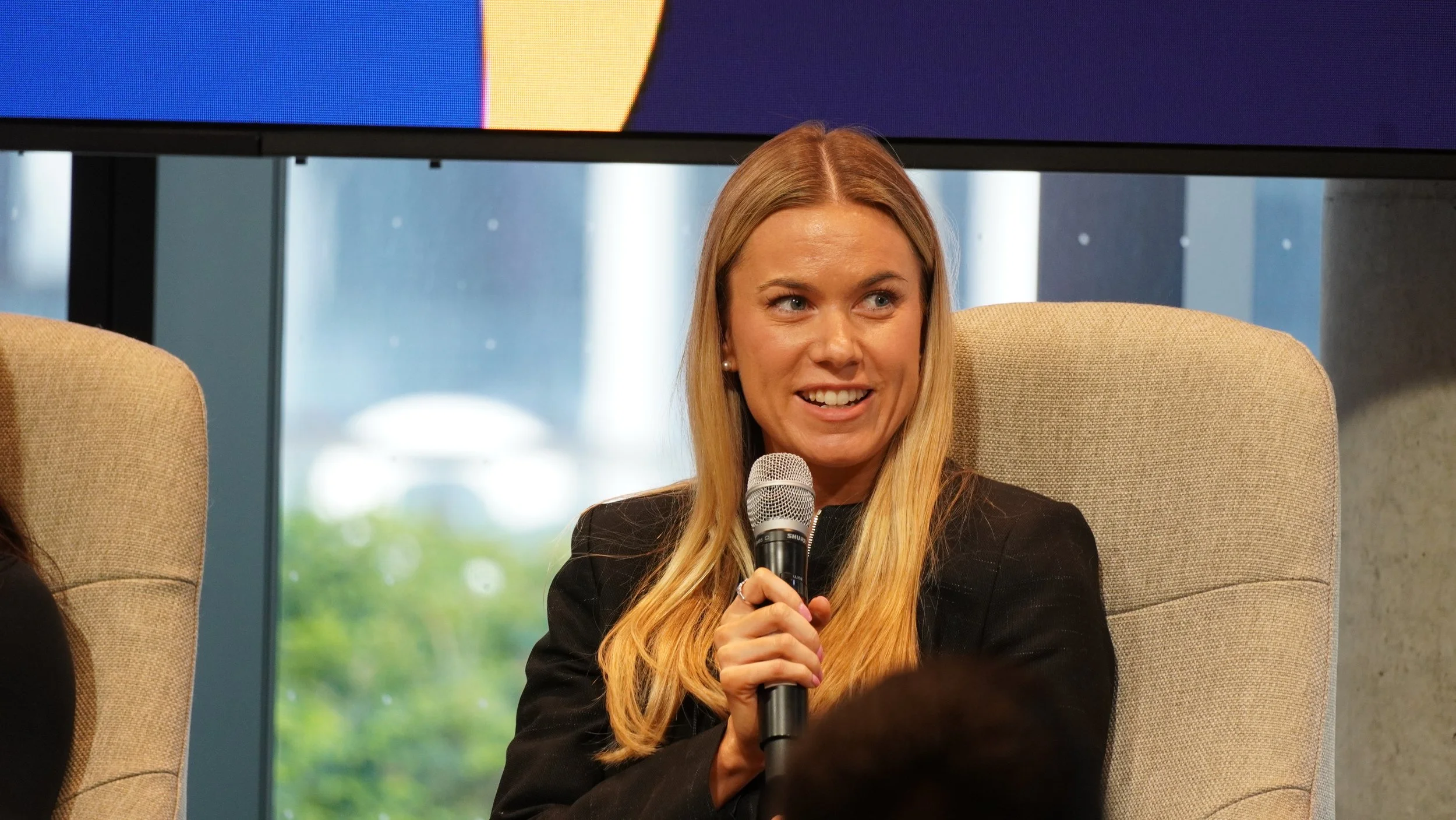 A woman with long blonde hair, wearing a black blazer, sitting in a beige armchair, holding a microphone, and speaking during an event or panel discussion.