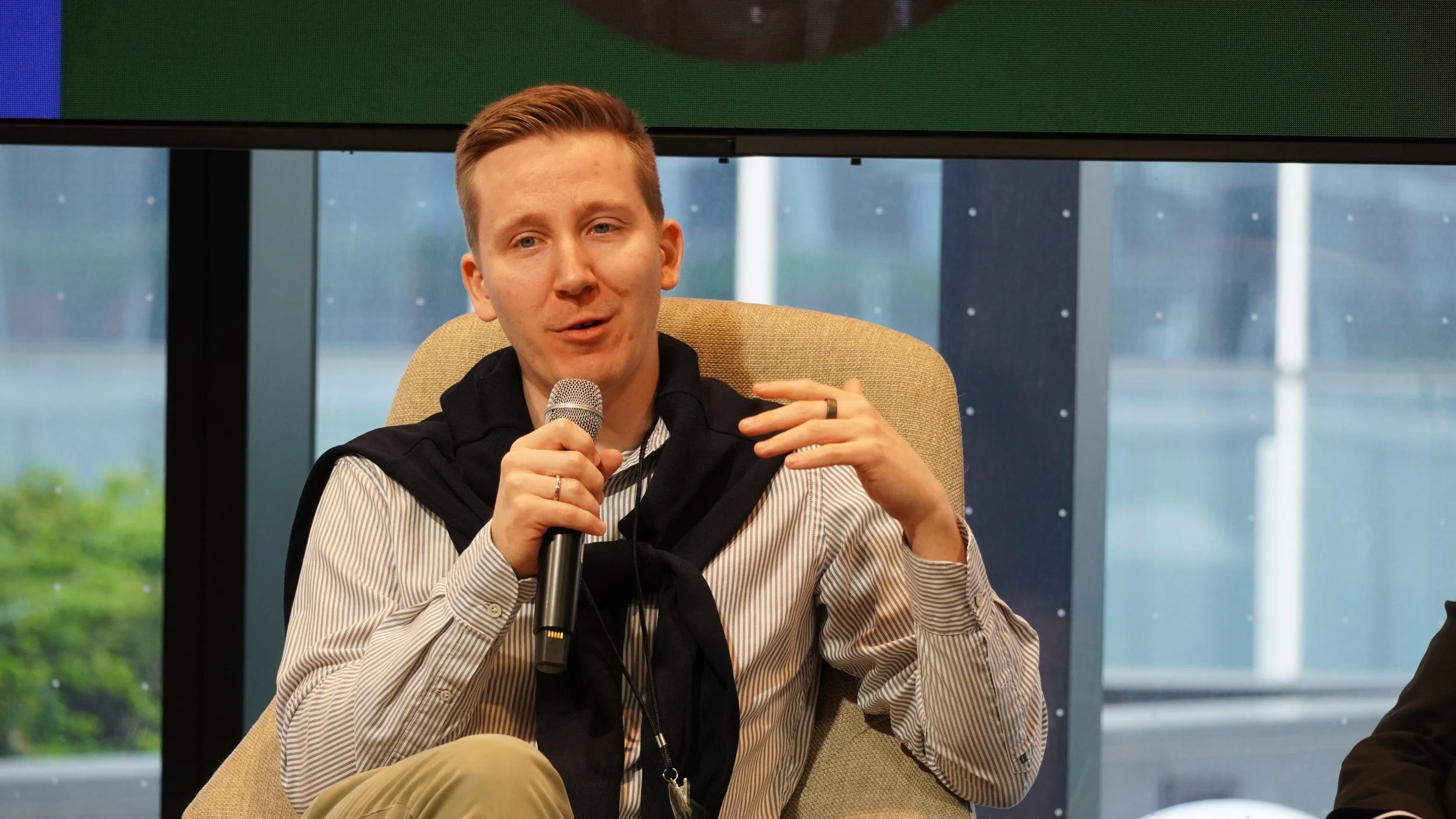 A man sitting on a beige chair, holding a microphone and speaking, with a large window showing a cityscape in the background.