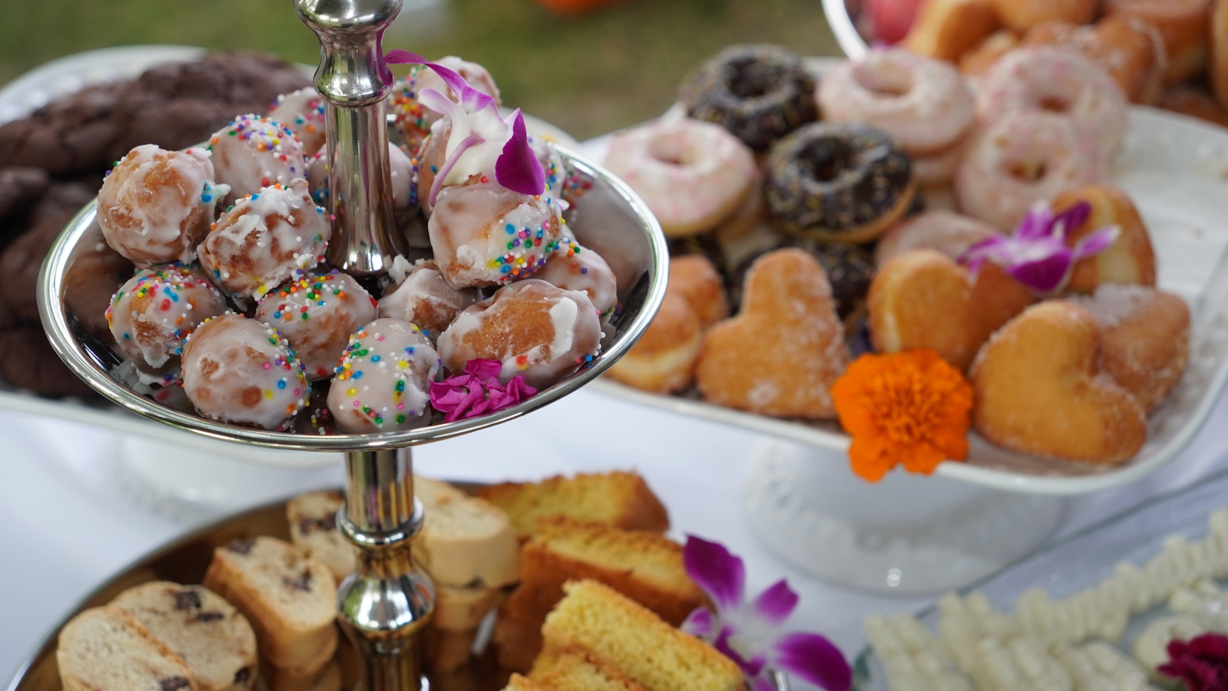 A silver tiered tray filled with assorted sweet treats, including donut holes with colorful sprinkles, cookies, and other confections, decorated with flowers.