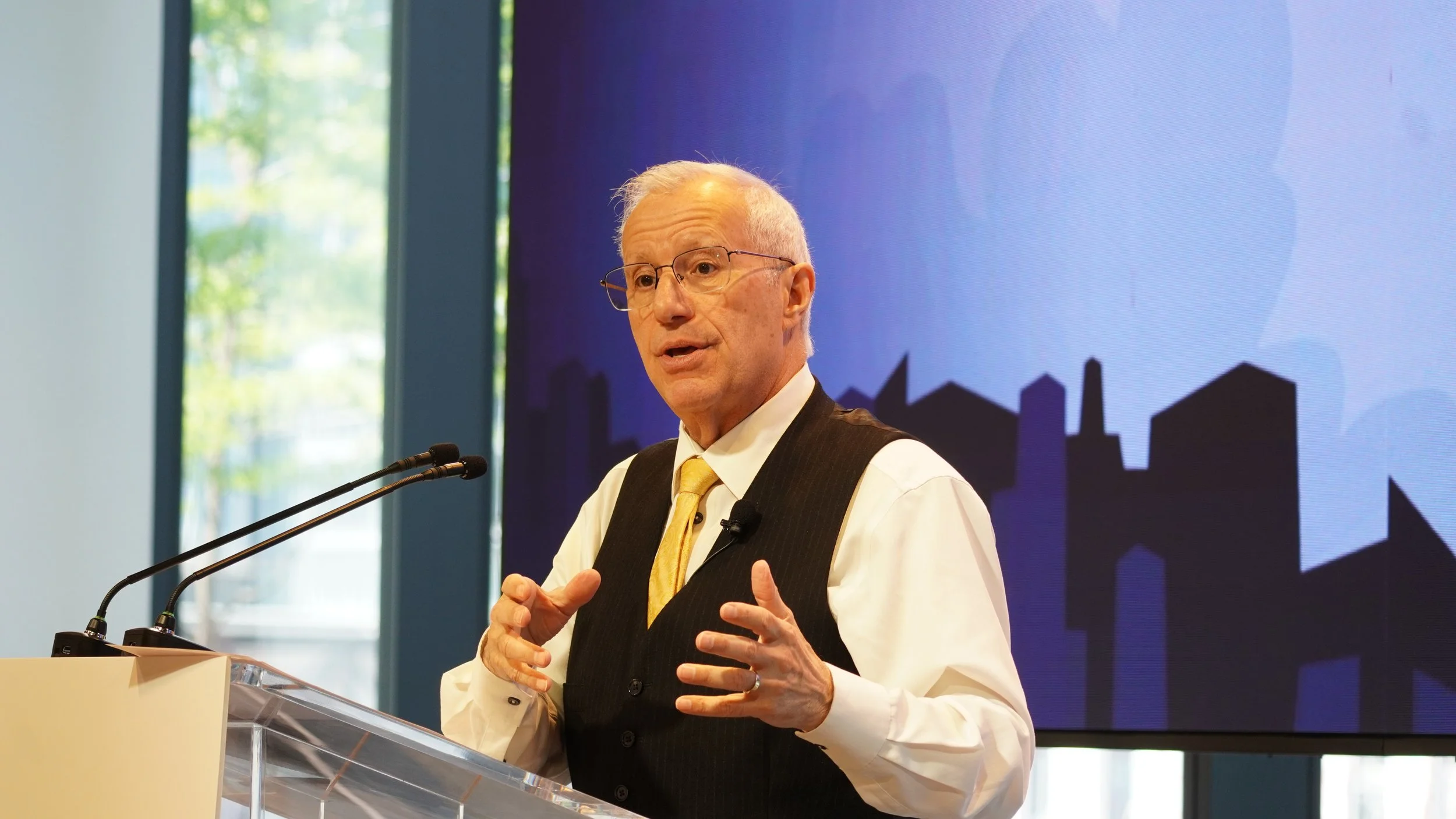 An older man with glasses and white hair giving a speech at a podium, wearing a white shirt, black vest, and yellow tie, with a city skyline and blue background on screen behind him.