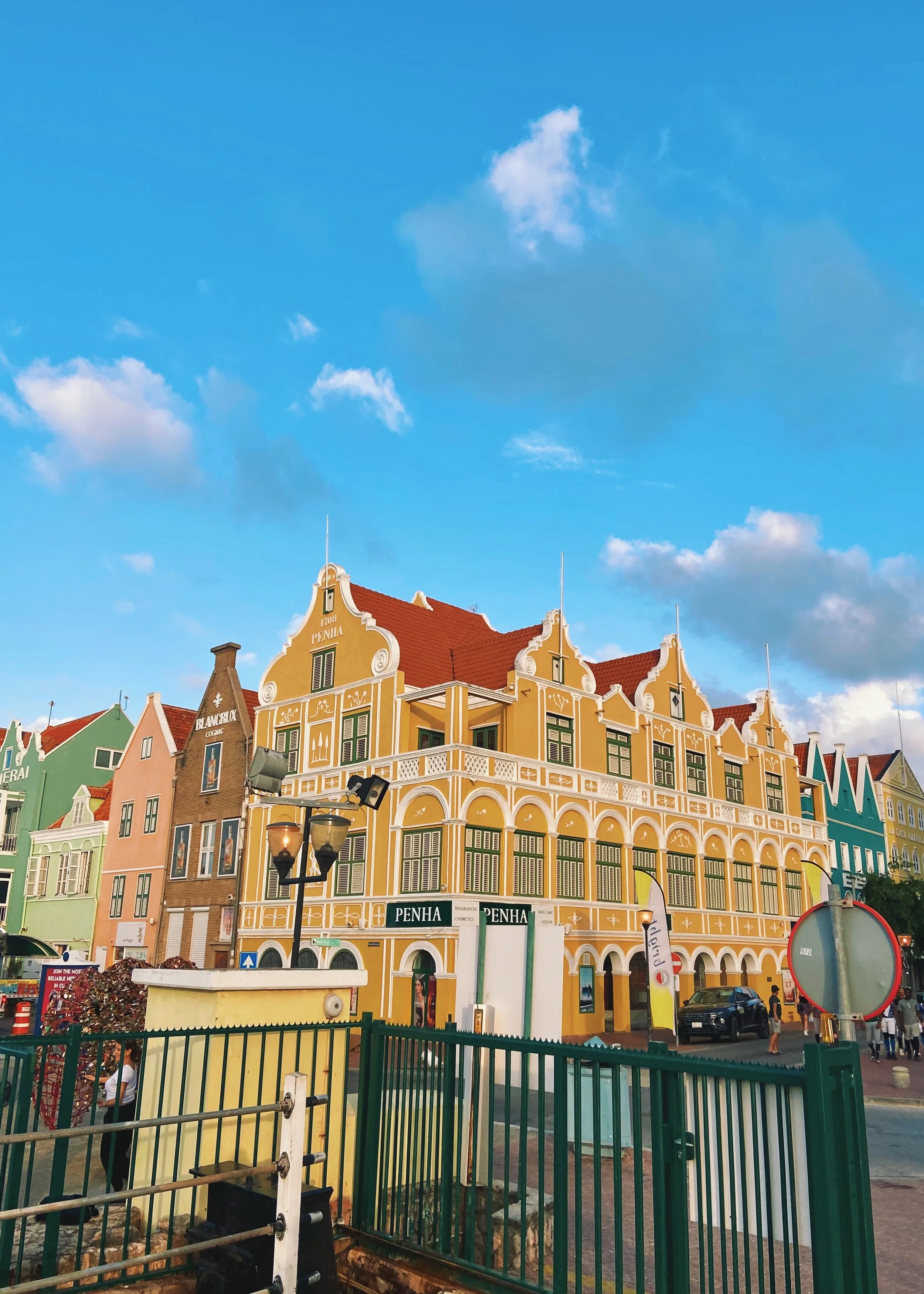 Colorful Dutch-style buildings on a street, with a blue sky and clouds overhead. The buildings are painted in yellow, green, pink, and blue, with decorative facades and red roofs.