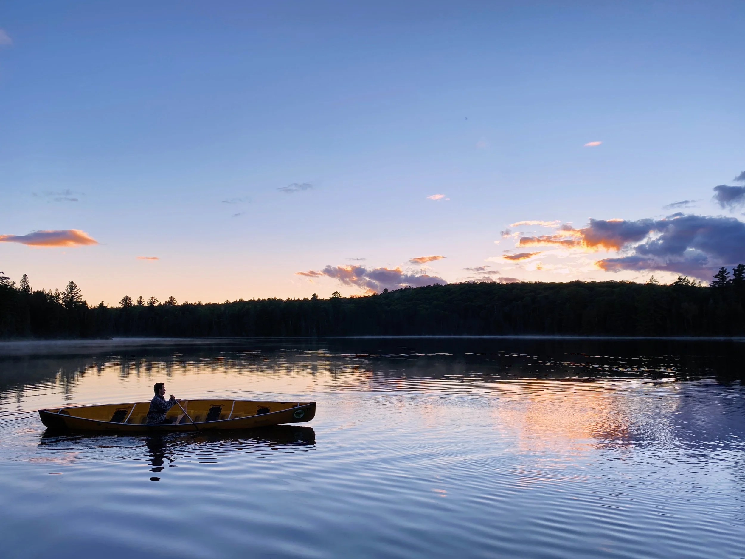 A person rowing a boat on a calm lake at sunset with a forested shoreline and a sky with scattered clouds reflected on the water.