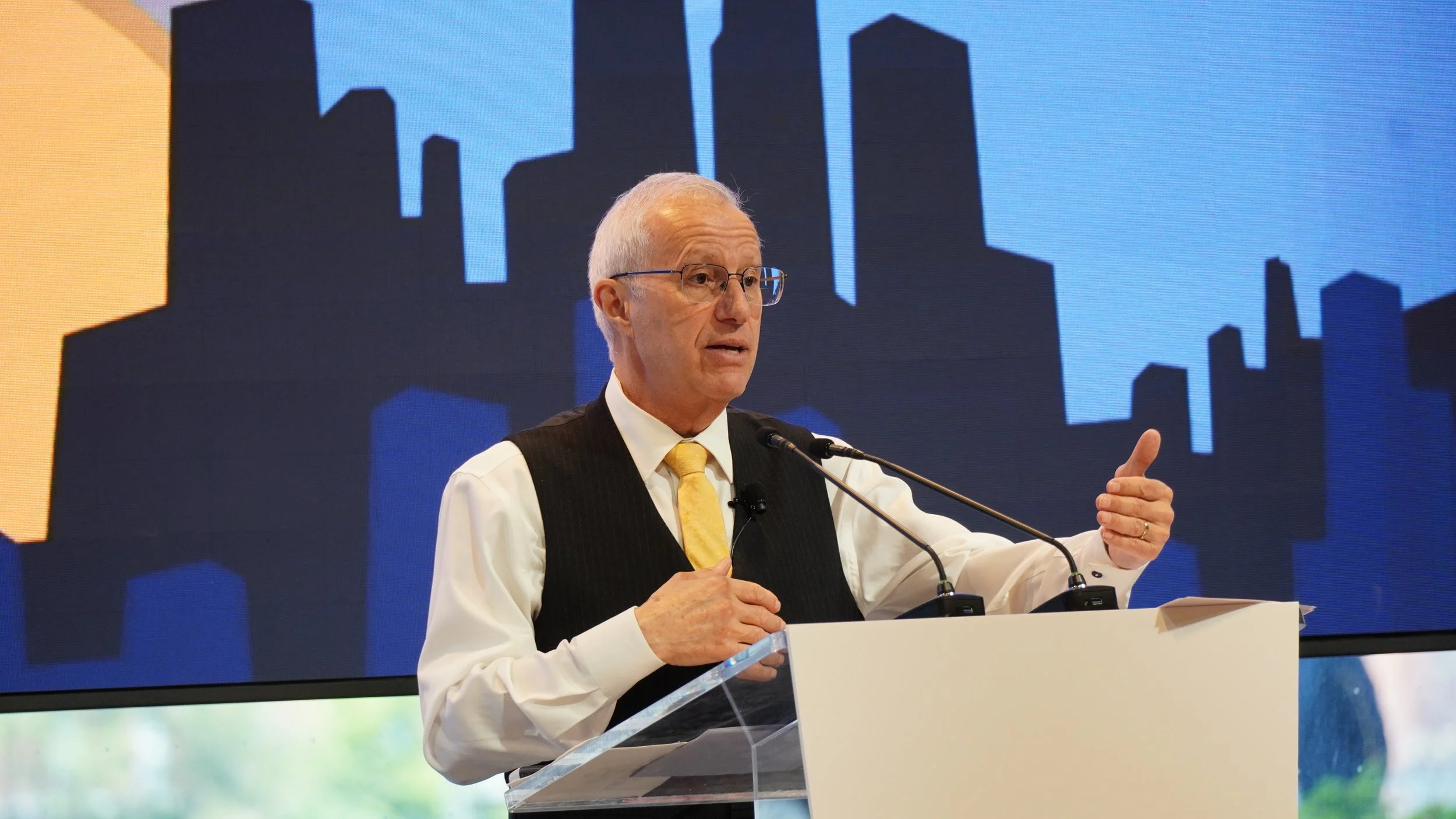 An older man with gray hair and glasses, wearing a white shirt, black vest, and yellow tie, speaking at a podium with a microphone. A large screen behind him displays a stylized city skyline in dark blue and black silhouettes.