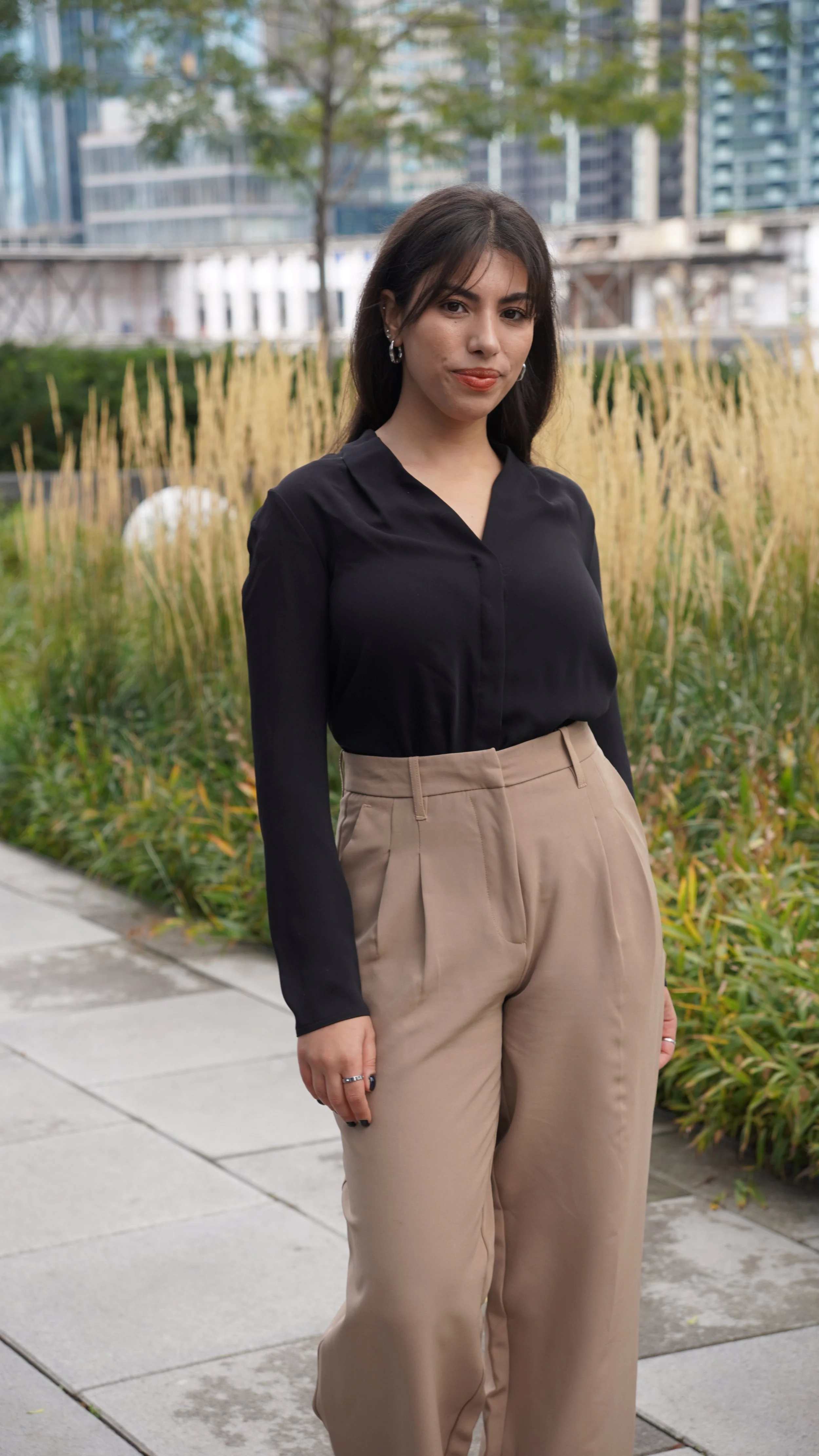 A young woman with dark hair, wearing a black blouse and beige pleated trousers, stands outdoors on a paved walkway with a cityscape and modern buildings in the background.