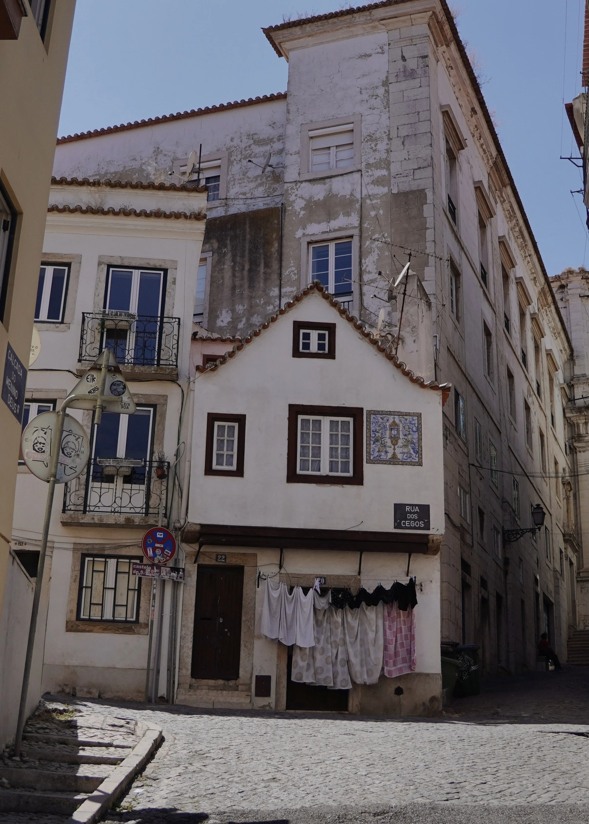 A small, white, house-like building with windows and laundry hanging outside, situated among taller, older buildings on a narrow cobblestone street in an urban setting.