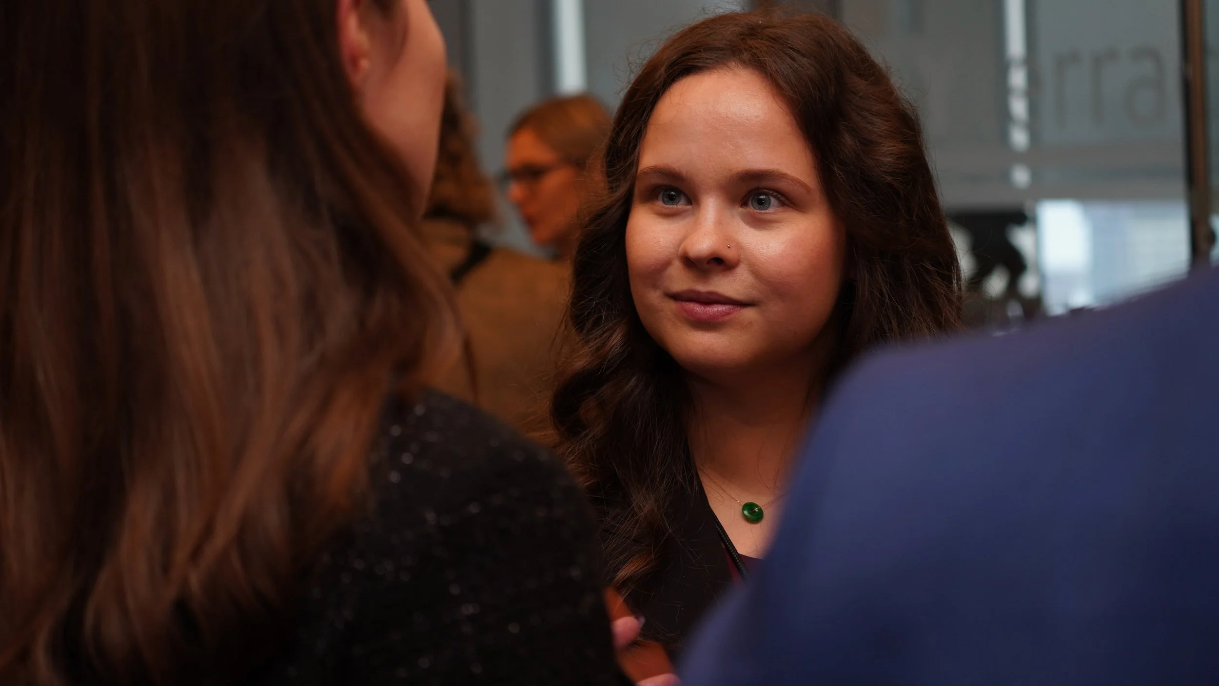 Young woman with brown hair and blue eyes listening to another person in a social setting.