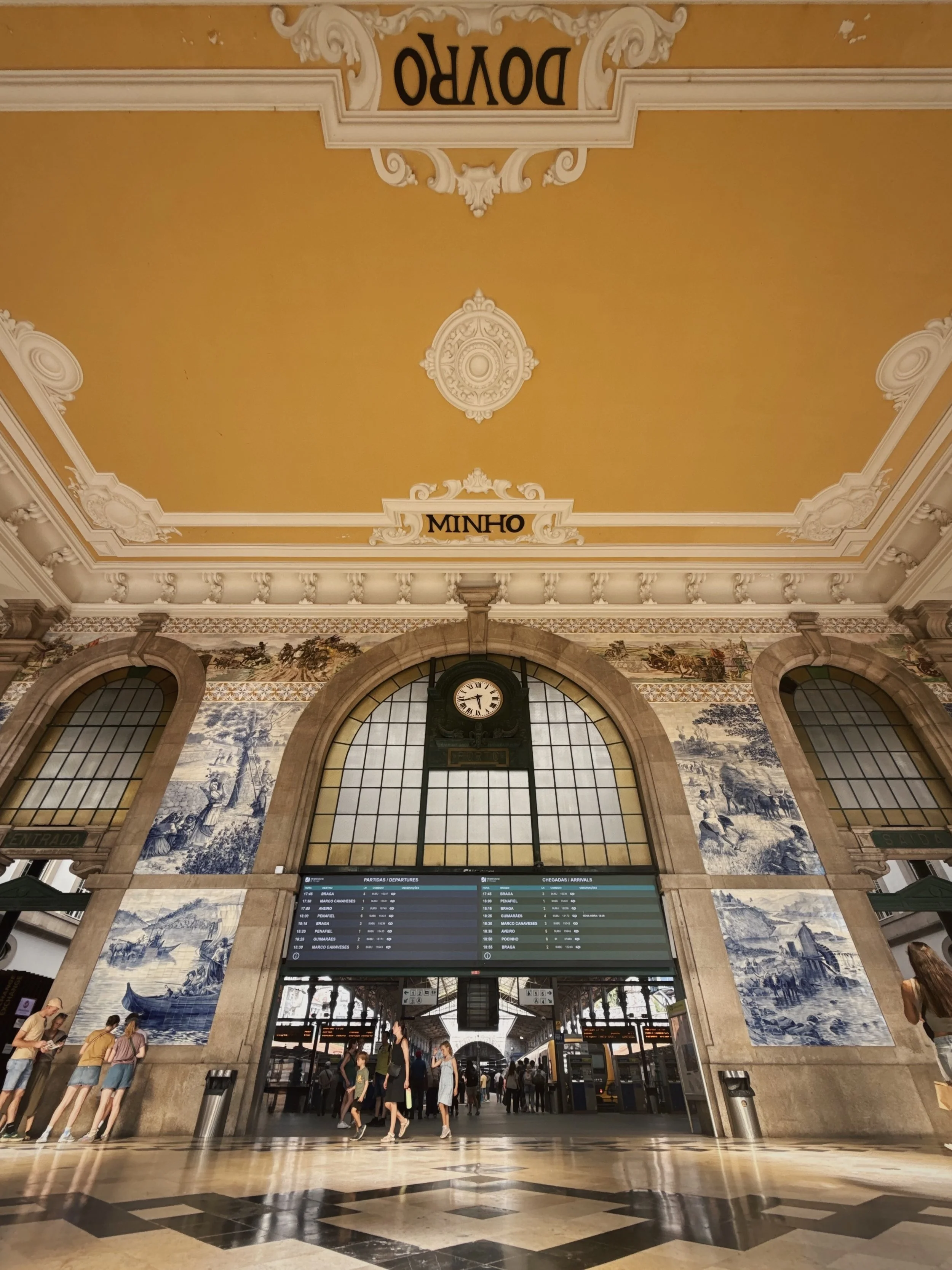 Interior of a historic train station with a large clock, departure and arrival screens, ornate architecture, blue and white tile murals, and travelers entering and exiting.