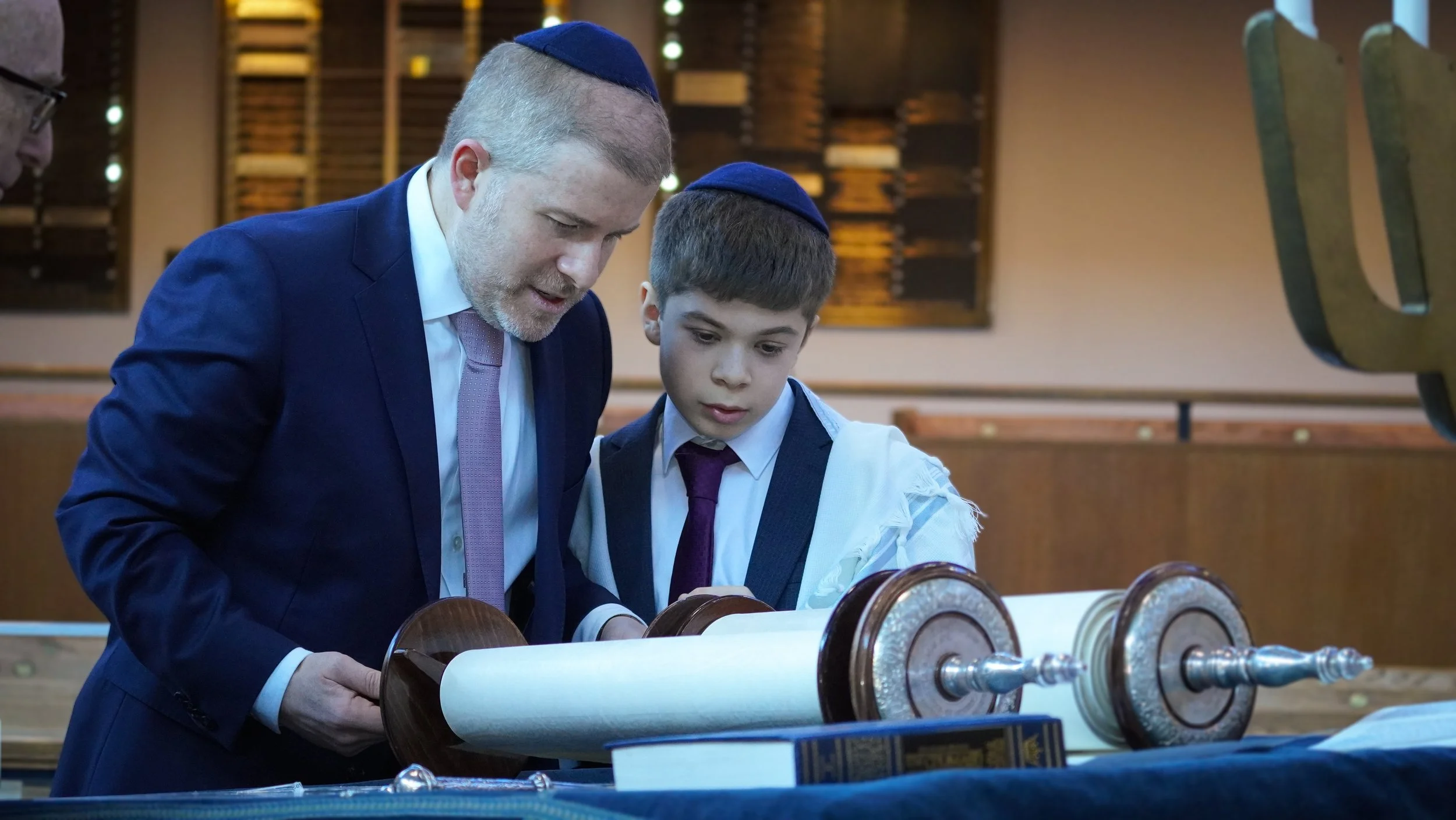 A man and a boy, both wearing kippahs, studying a Torah scroll in a synagogue. The man is dressed in a dark suit and tie, and the boy is in a white shirt with a dark vest and tie.