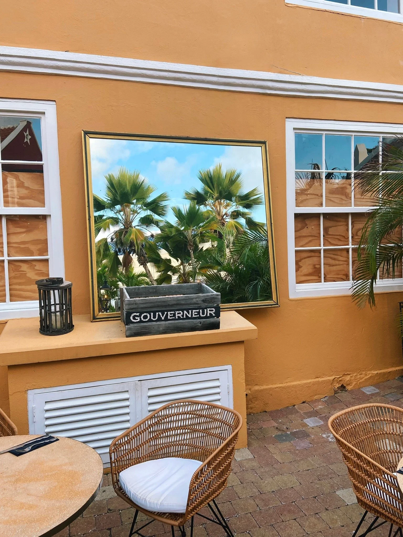 A mirror reflecting palm trees and sky, placed on a yellow wall beside windows, with a wooden crate labeled 'GOUVERNEUR' and a candleholder on a ledge. Wicker chairs and a round table are in the foreground.