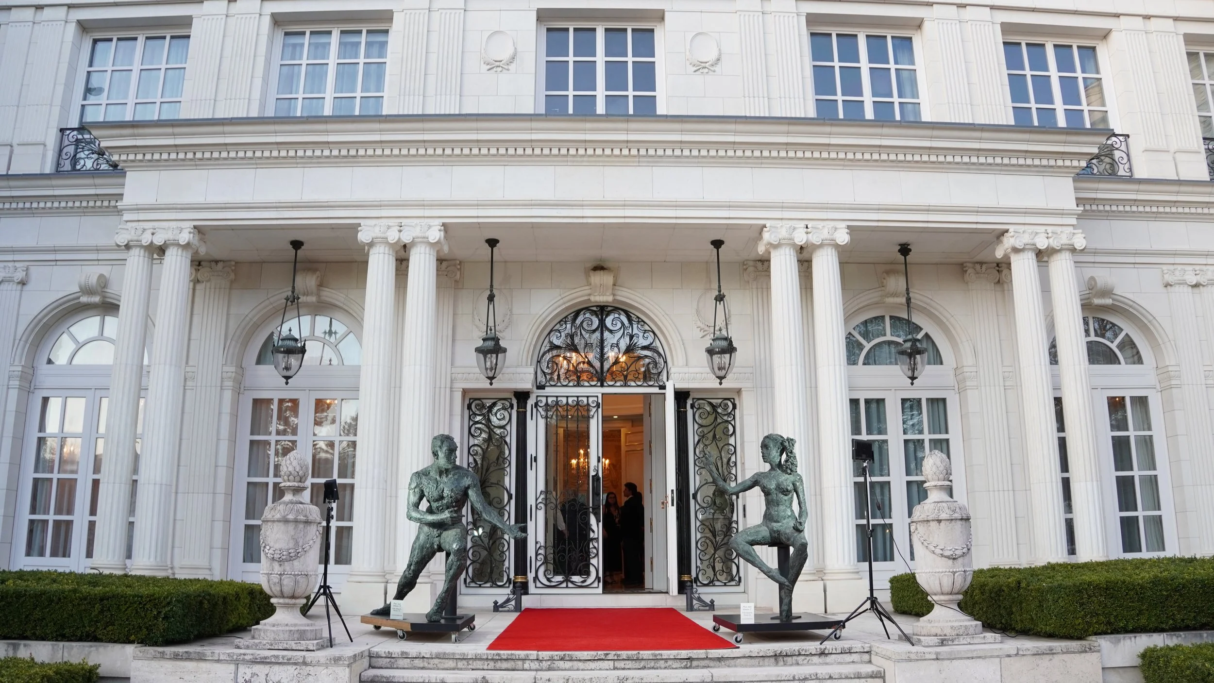 The entrance of a grand, white, neoclassical building with tall columns, decorative iron gate, and two sculptures of intertwined male and female figures on either side. A red carpet leads to the open door, with greenery bordering the stairs.
