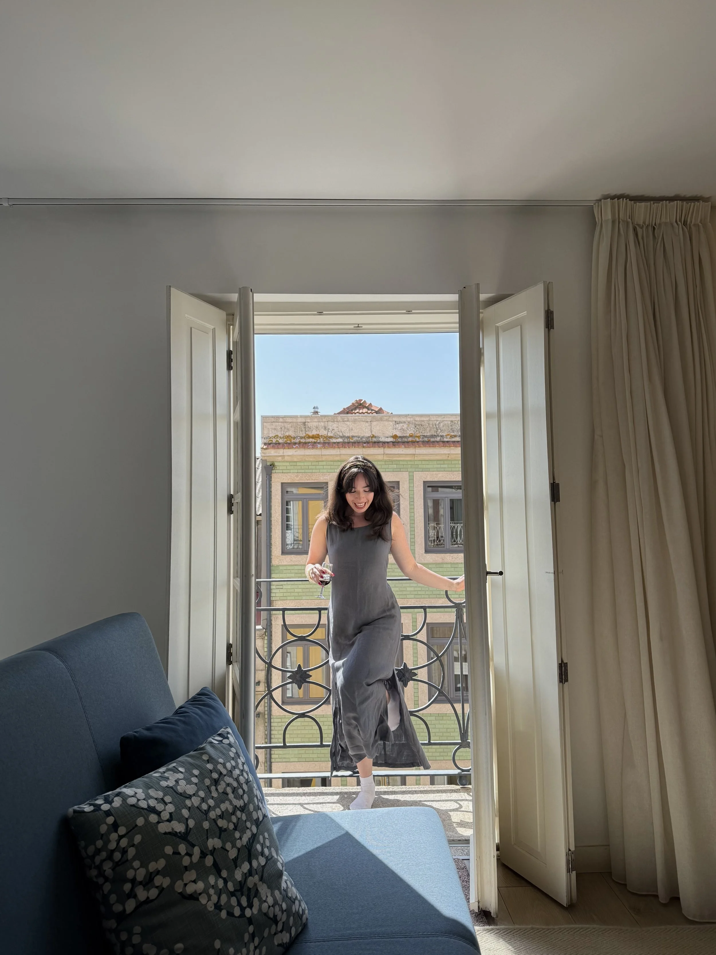 A woman in a gray dress is coming into a room through a balcony door, holding a glass of wine, with sunlight streaming in.