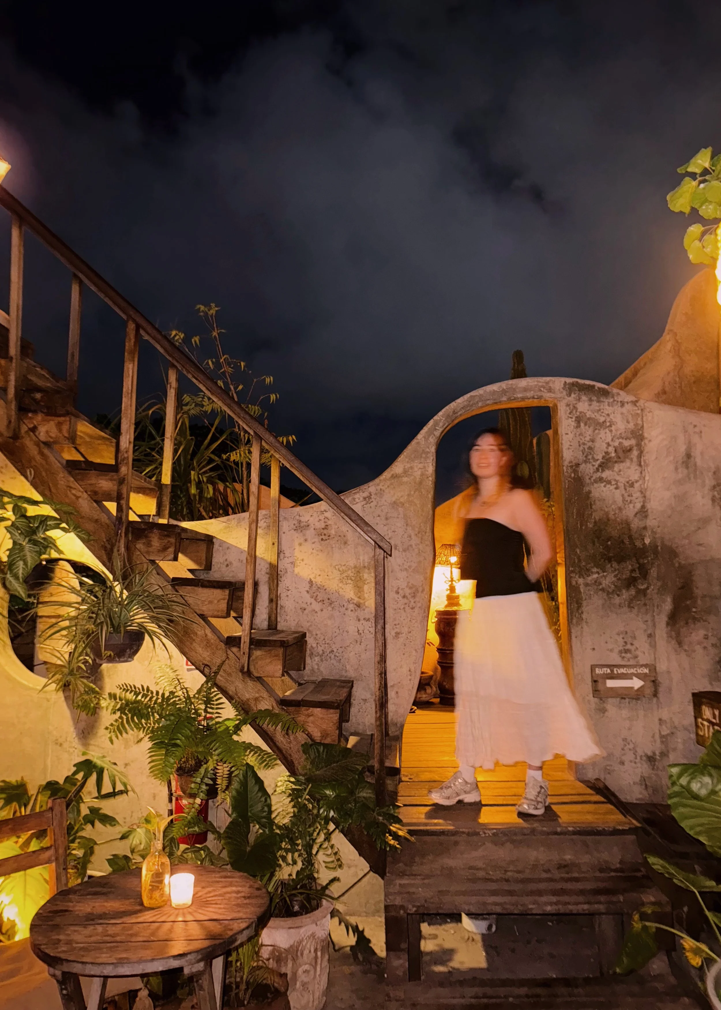 A woman in a black top and white skirt standing on a wooden porch at night, surrounded by plants and a lit lamp inside, with dark clouds in the sky.