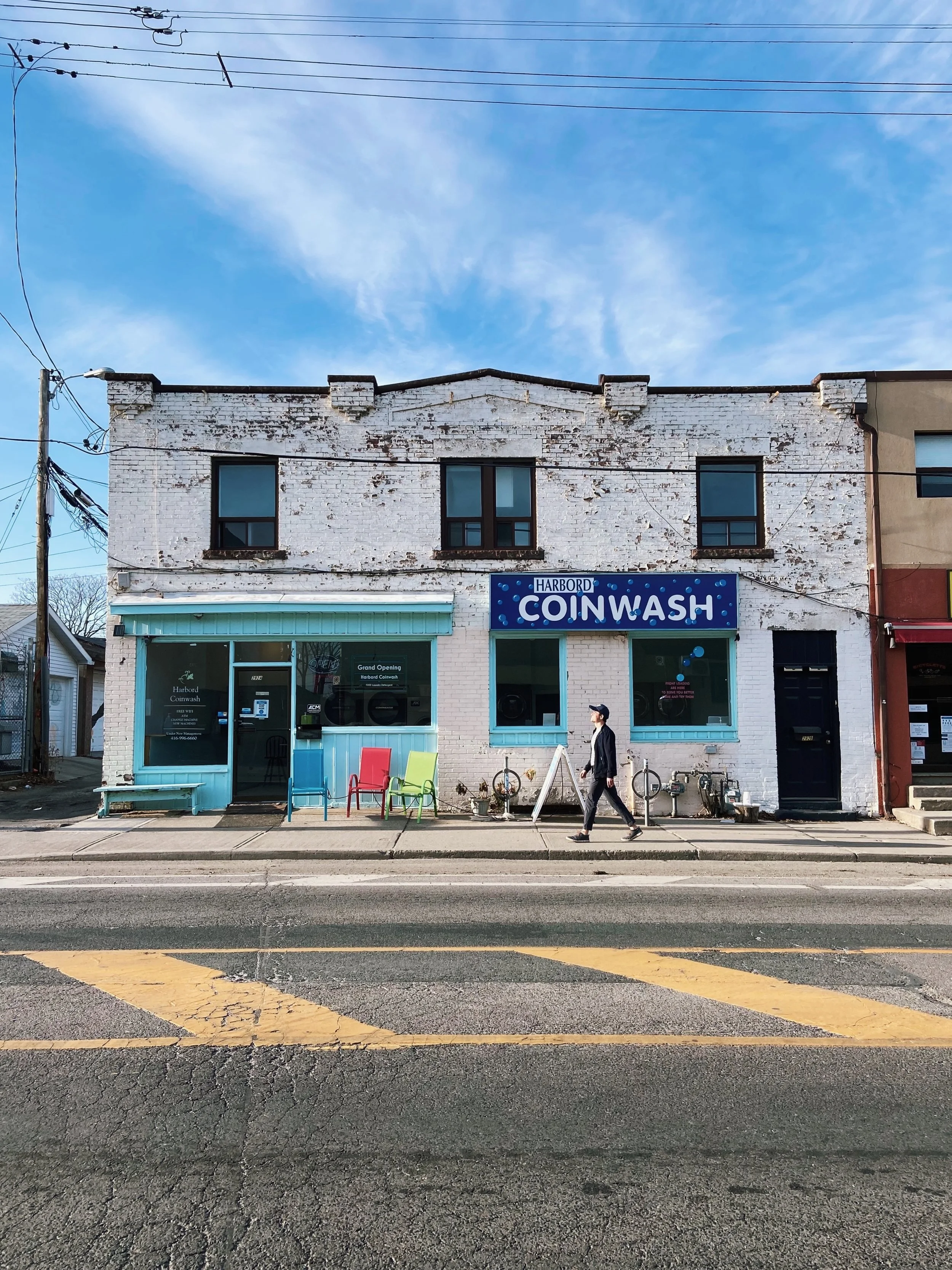 A two-story white brick building with a sign that reads 'Harbord Coinwash' in blue letters, with smaller text indicating grand opening, and colorful chairs outside. A person is walking on the sidewalk in front of the building.