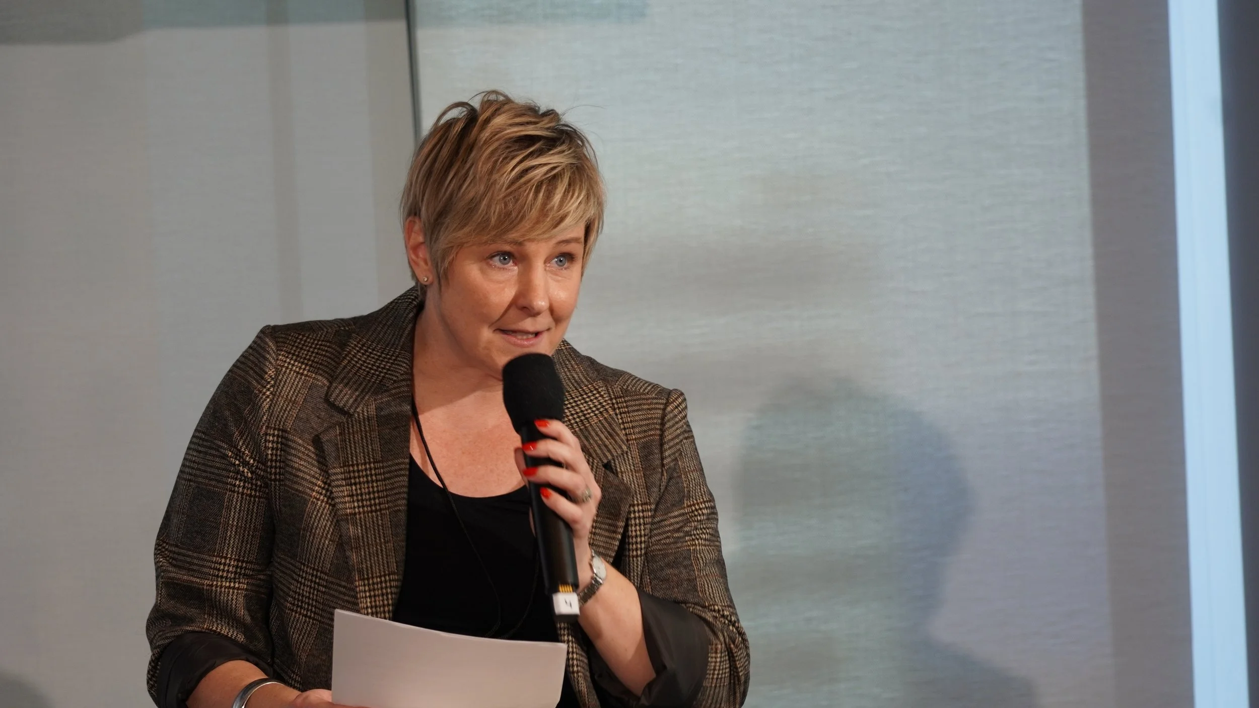 A woman with short blonde hair, wearing a checkered blazer and black top, speaking into a microphone while holding a piece of paper.