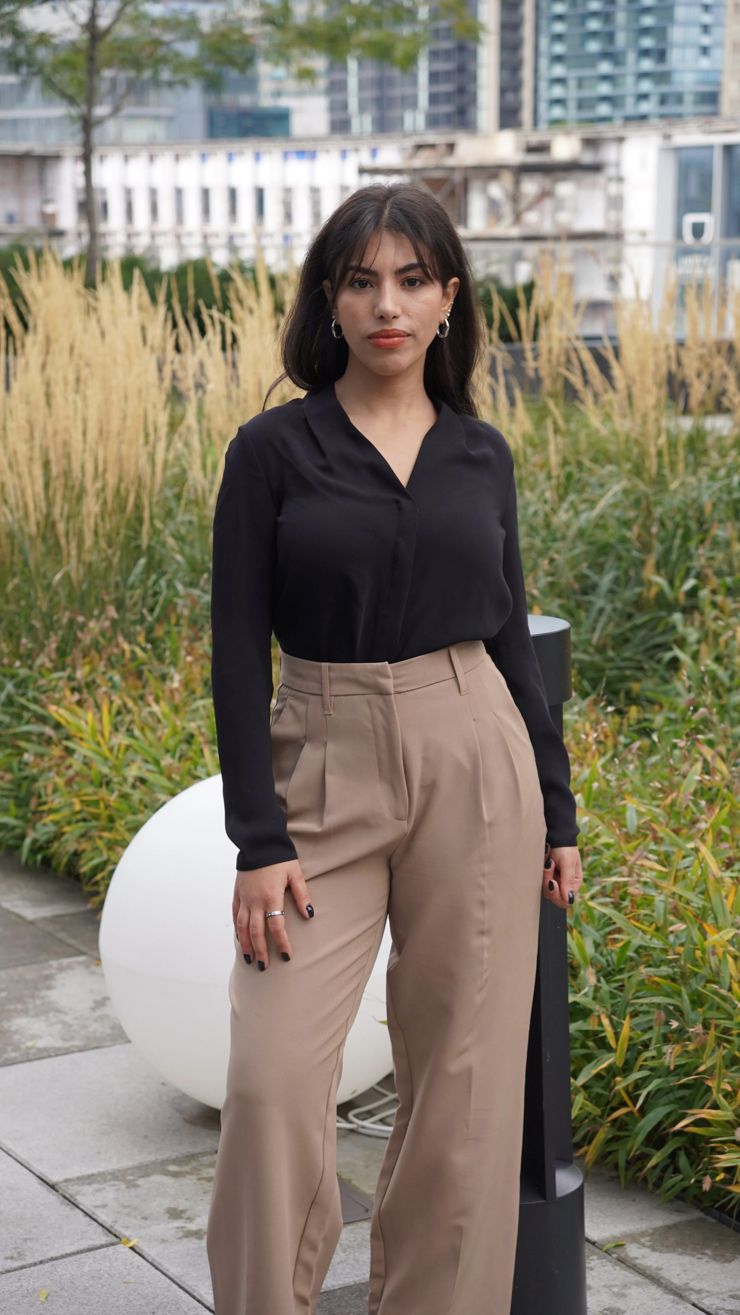 A young woman with dark hair and hoop earrings standing outdoors in a city park with green plants and tall buildings in the background, wearing a black blouse and beige wide-leg trousers.