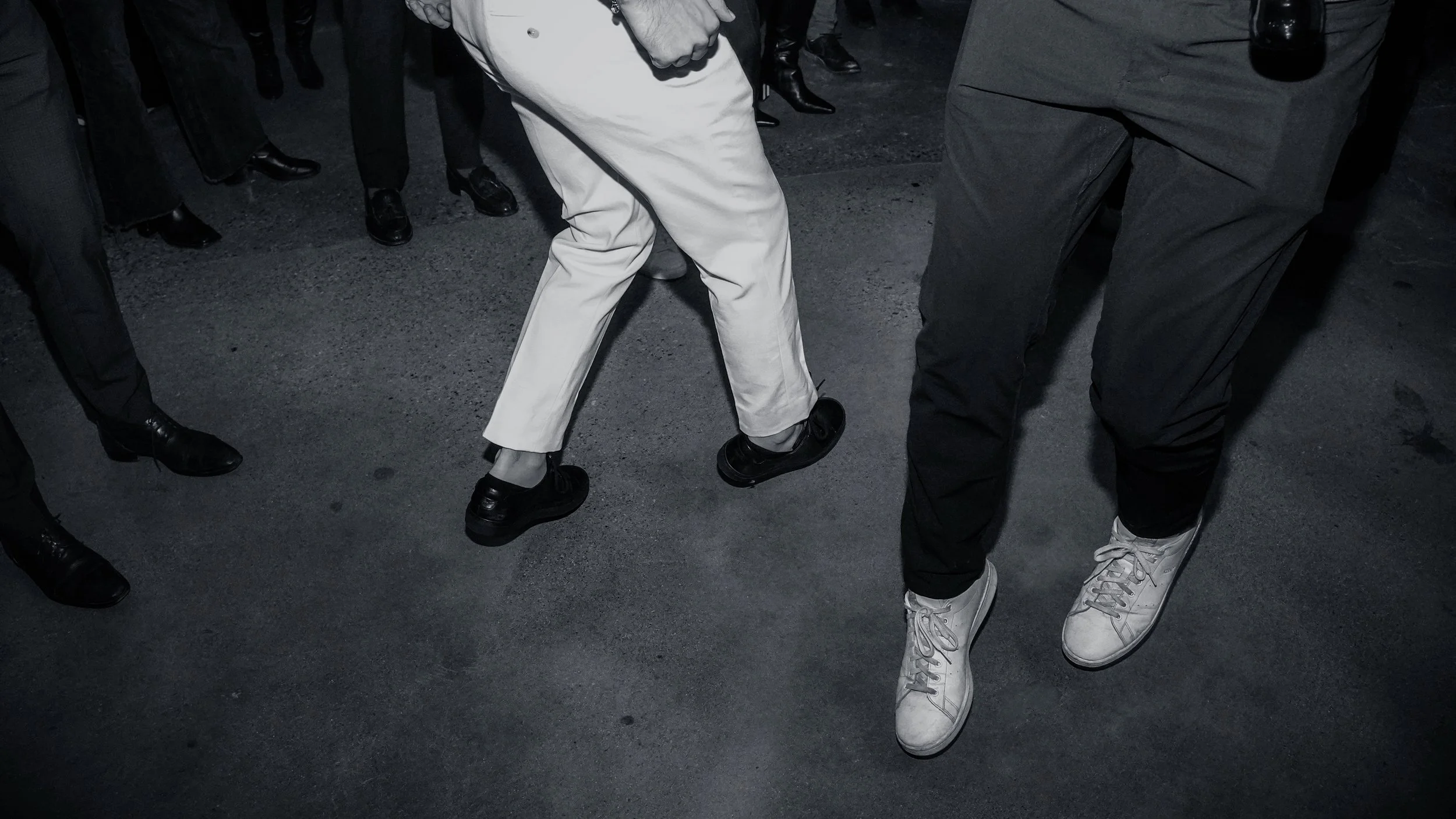 People standing on a dark street at night, wearing dark clothing and shoes, with one person in white pants and white sneakers, and another in black pants and shoes.