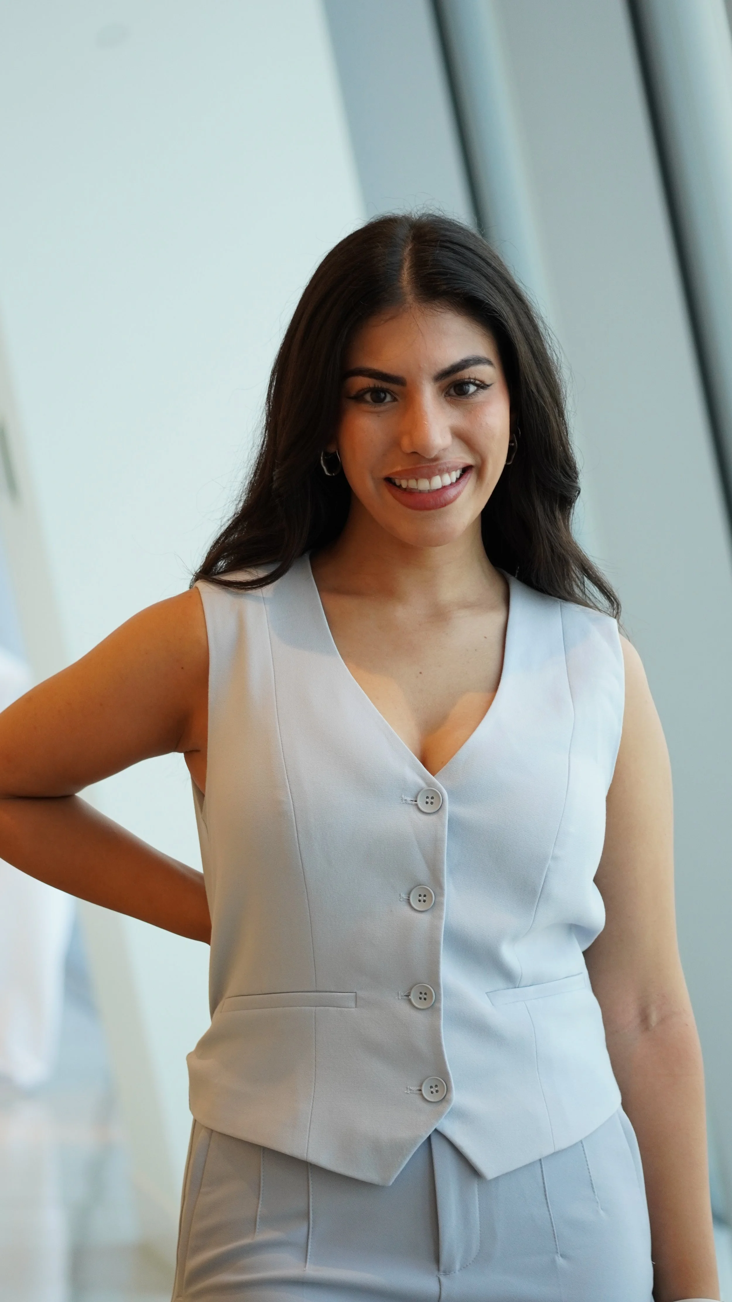 A young woman with long dark hair and earrings, smiling, wearing a light-colored sleeveless top with buttons, standing indoors near large windows.