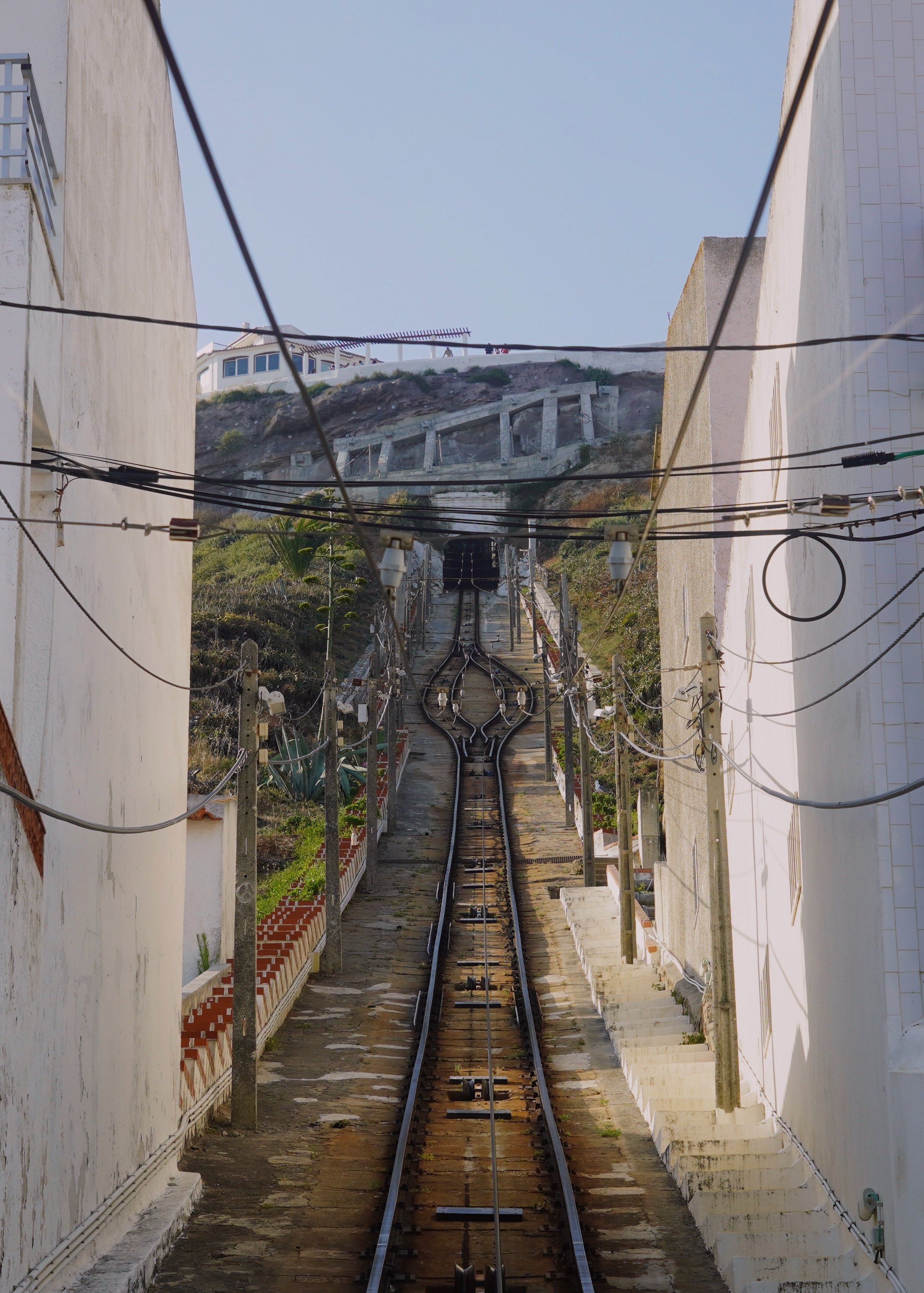 Railway tracks descending between buildings, with electrical wires overhead and a hillside in the background.