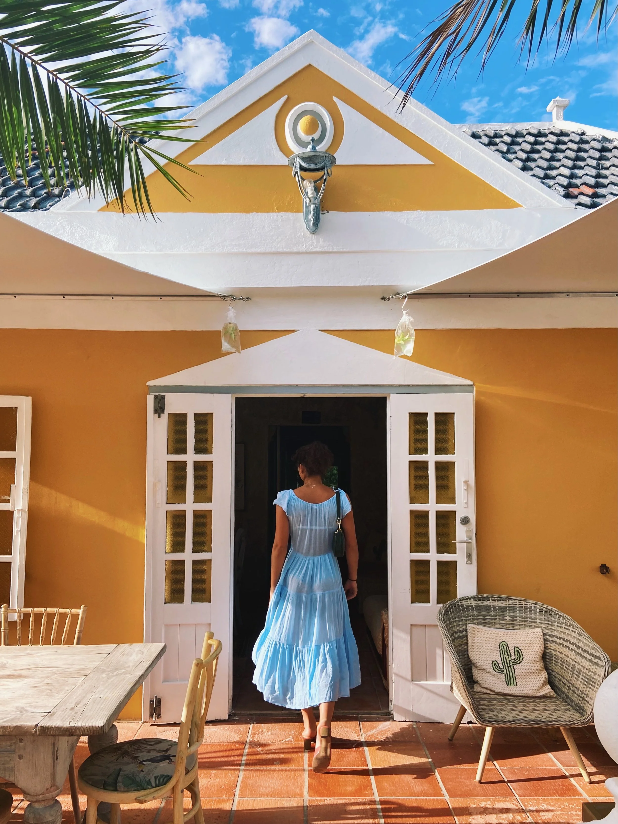 A woman in a light blue dress with a black purse walking into a yellow home with white double doors, outdoor furniture including a wooden table, chairs, a cushioned wicker chair with a cactus pillow, and stringed lights, under a blue sky with scatter