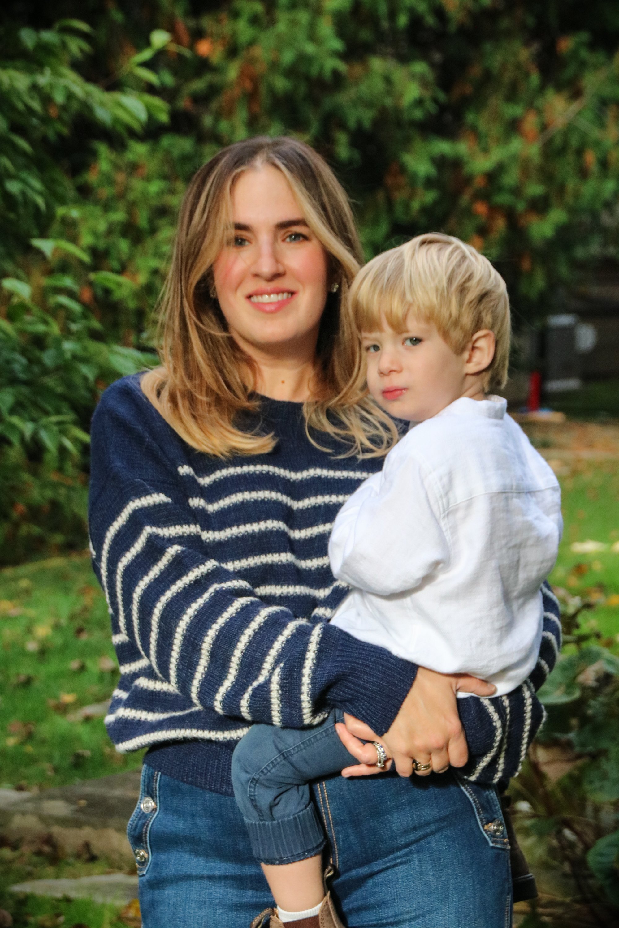 A woman with shoulder-length light brown hair, wearing a navy blue sweater with white stripes, holding a young blond-haired boy in a white shirt in an outdoor garden setting.