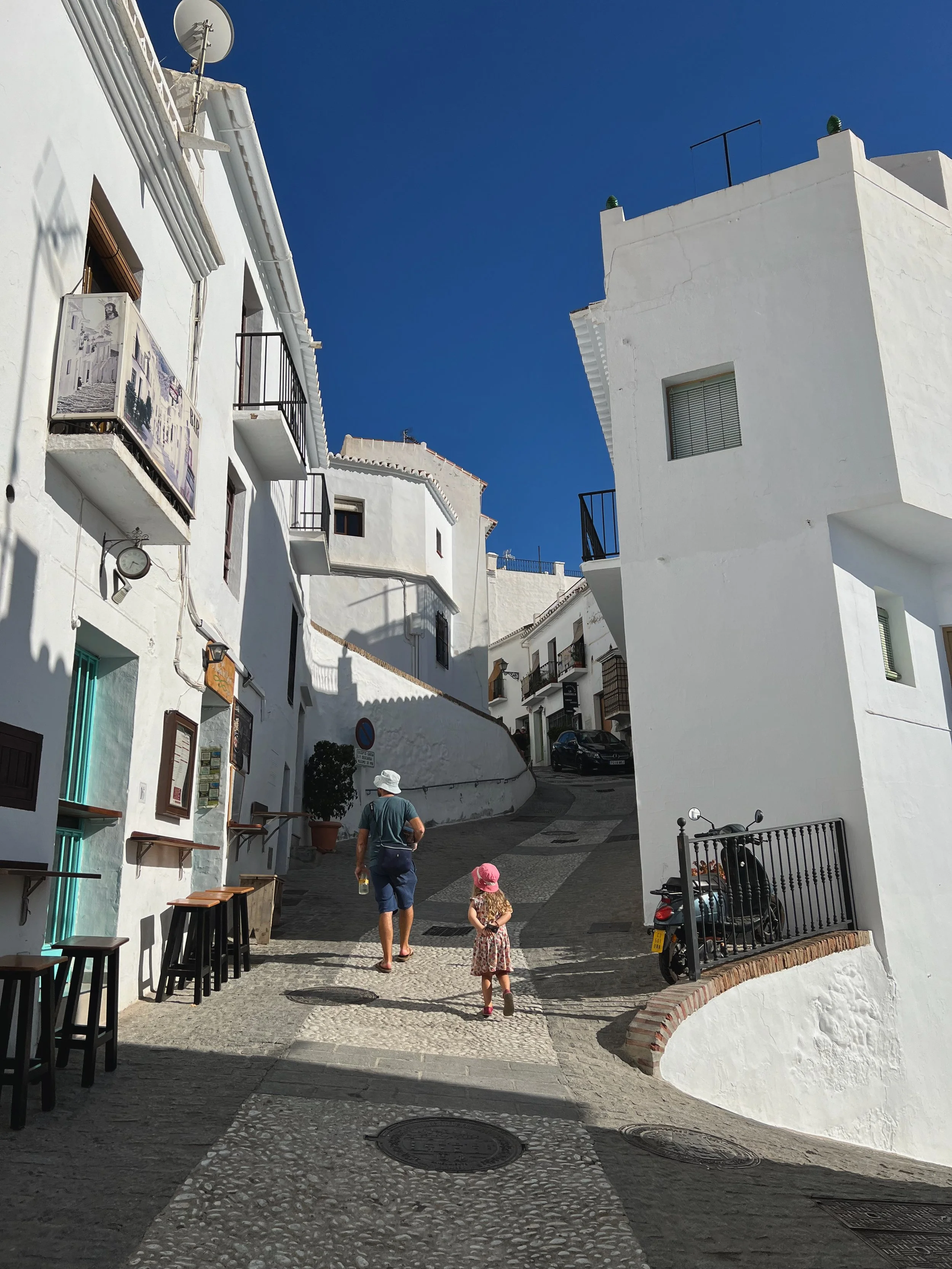 A man and a young girl walk up a narrow cobblestone street with white buildings on either side, some with small balconies and potted plants. The sky is clear and blue.
