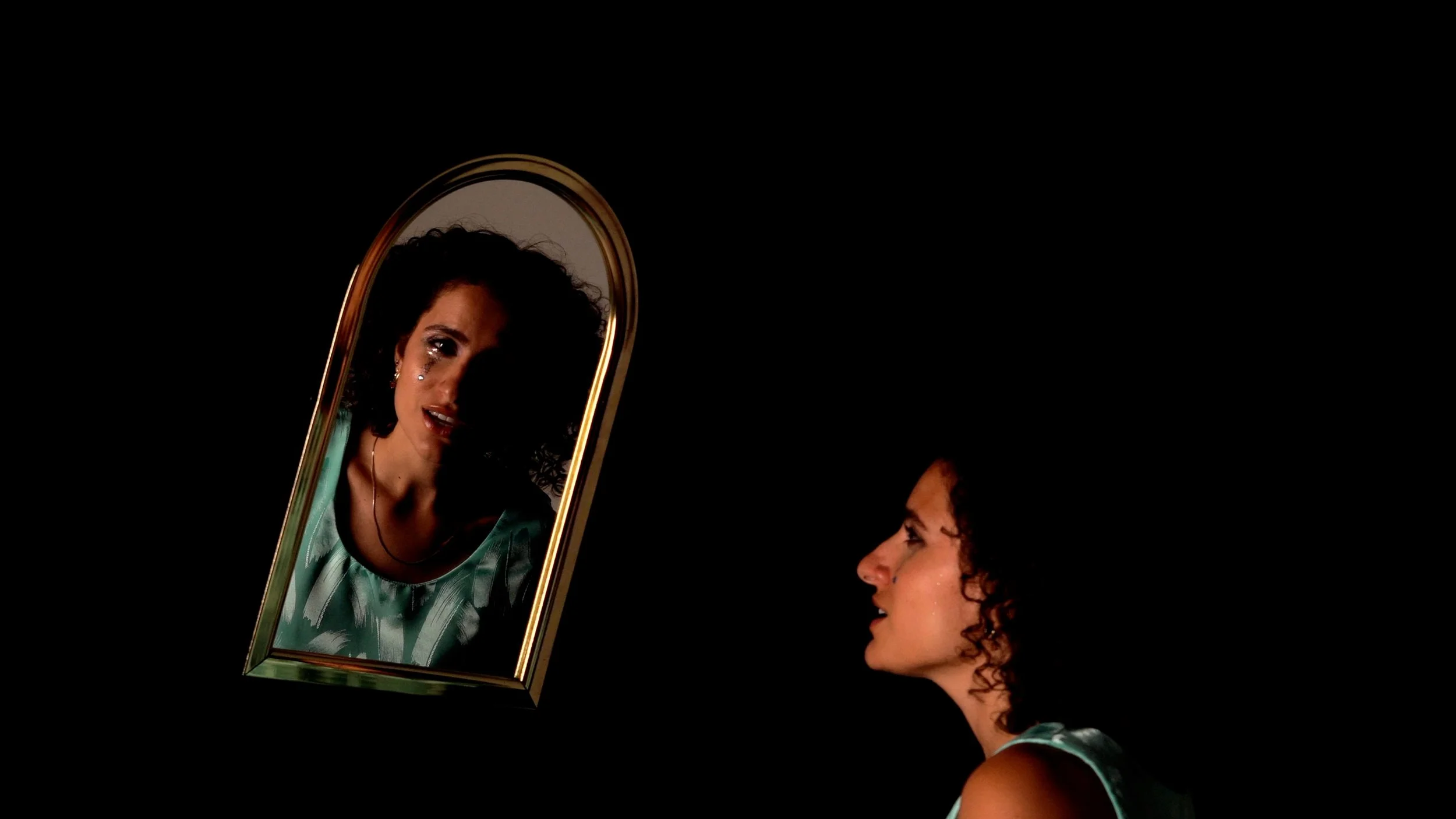 A woman with dark curly hair and shiny makeup looks at her reflection in a framed mirror against a black background.