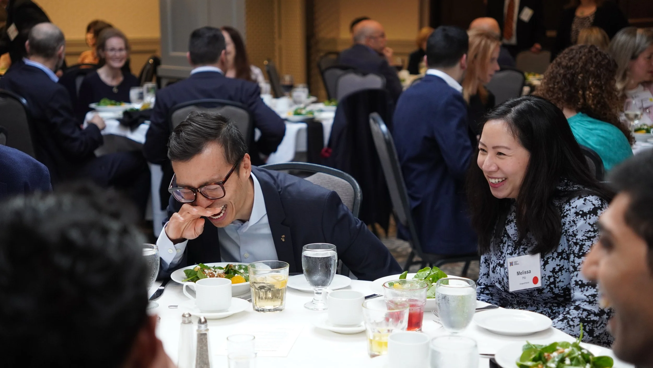 People laughing and enjoying a meal at a formal event or conference, seated around a table with plates of salad, drinks, and tableware.