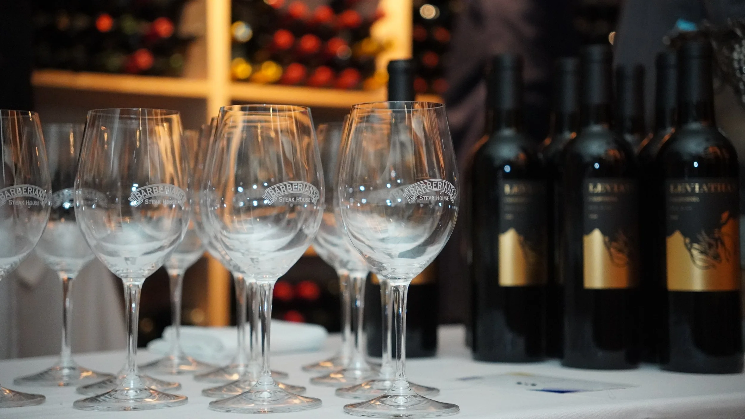Several empty wine glasses with 'Barberians Steak House' engraved on them, arranged on a table, with bottles of wine or spirits behind them, in a dimly lit setting.