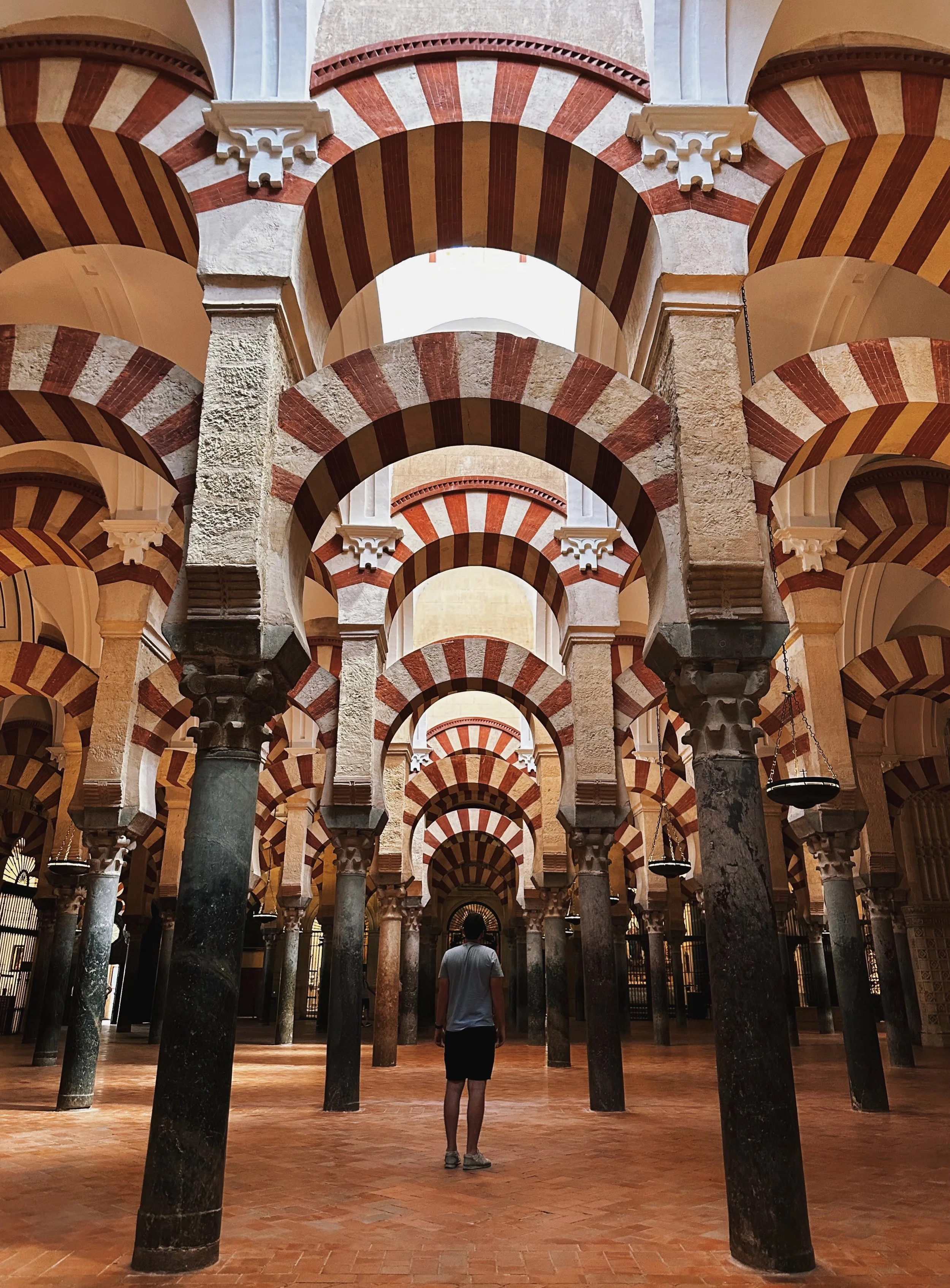 Interior view of the Great Mosque-Cathedral of Córdoba with multiple red and white striped arches and columns, a person walking underneath.