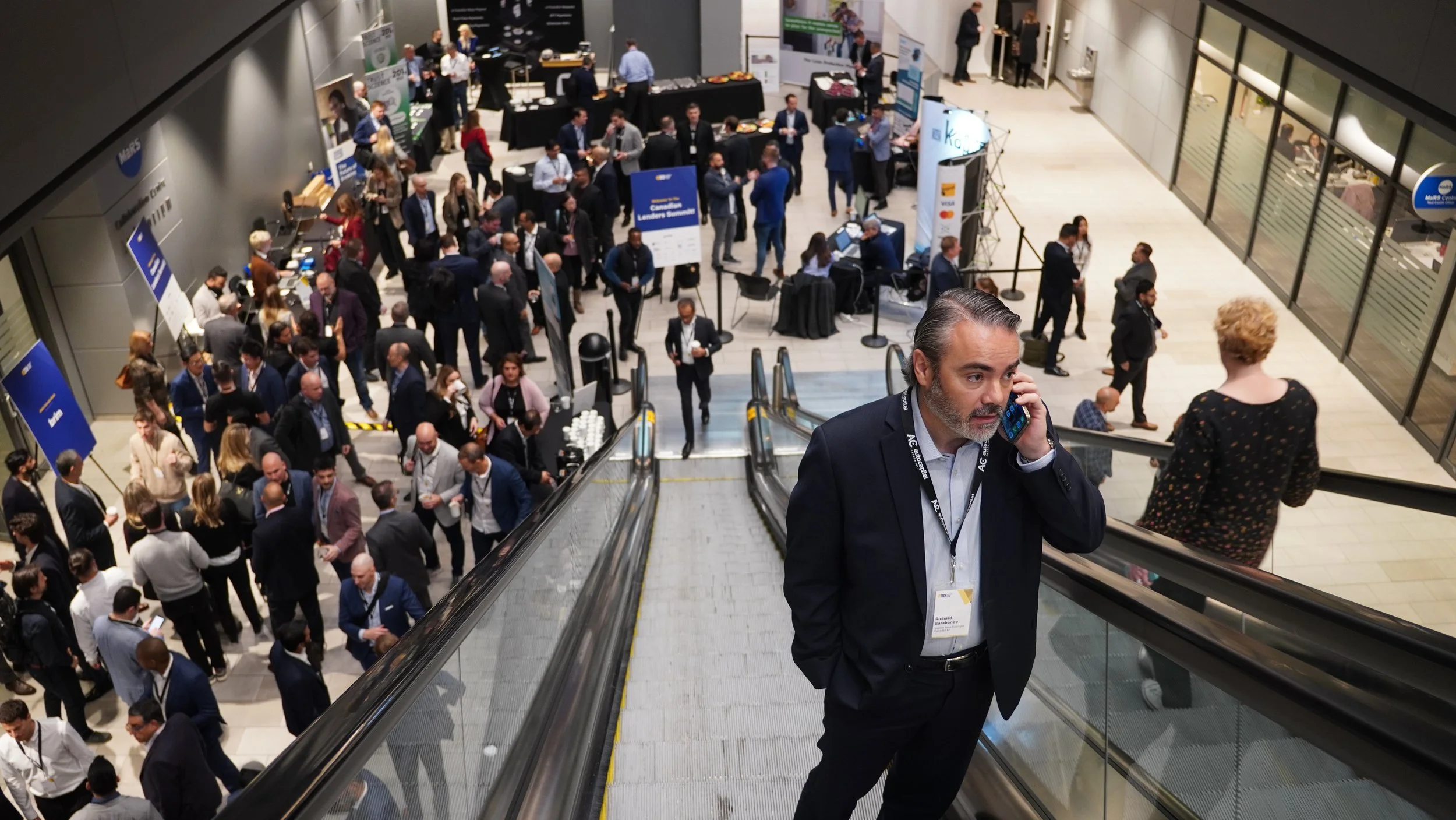 A crowded conference or trade show with many people walking, talking, and standing near booths and displays. A man in a suit is on an escalator talking on his phone, and a woman with curly blonde hair is also on the escalator facing away.