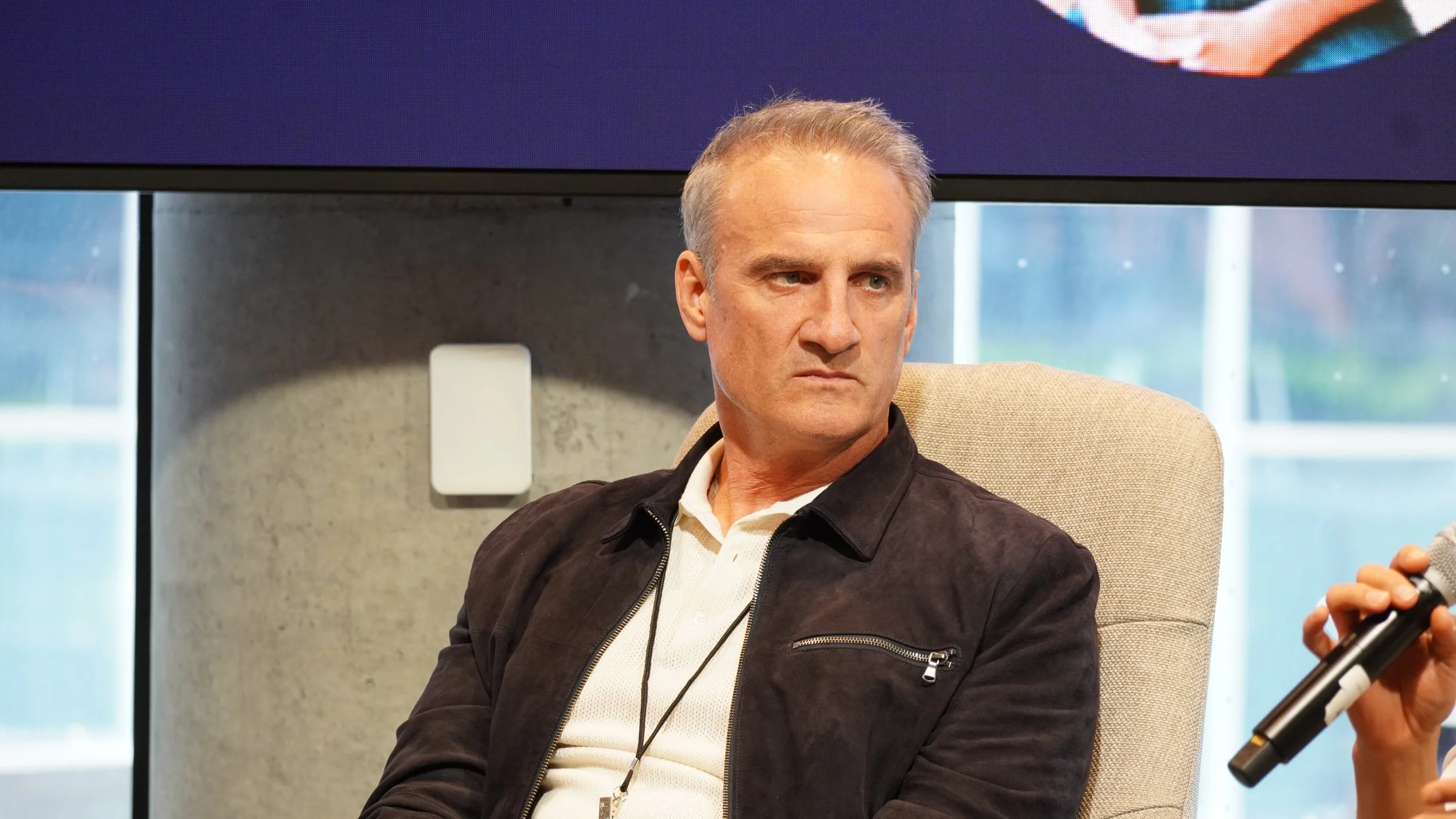 A man with gray hair staring intently while seated in a beige chair, with a gray wall and windows in the background. Someone nearby is holding a microphone.