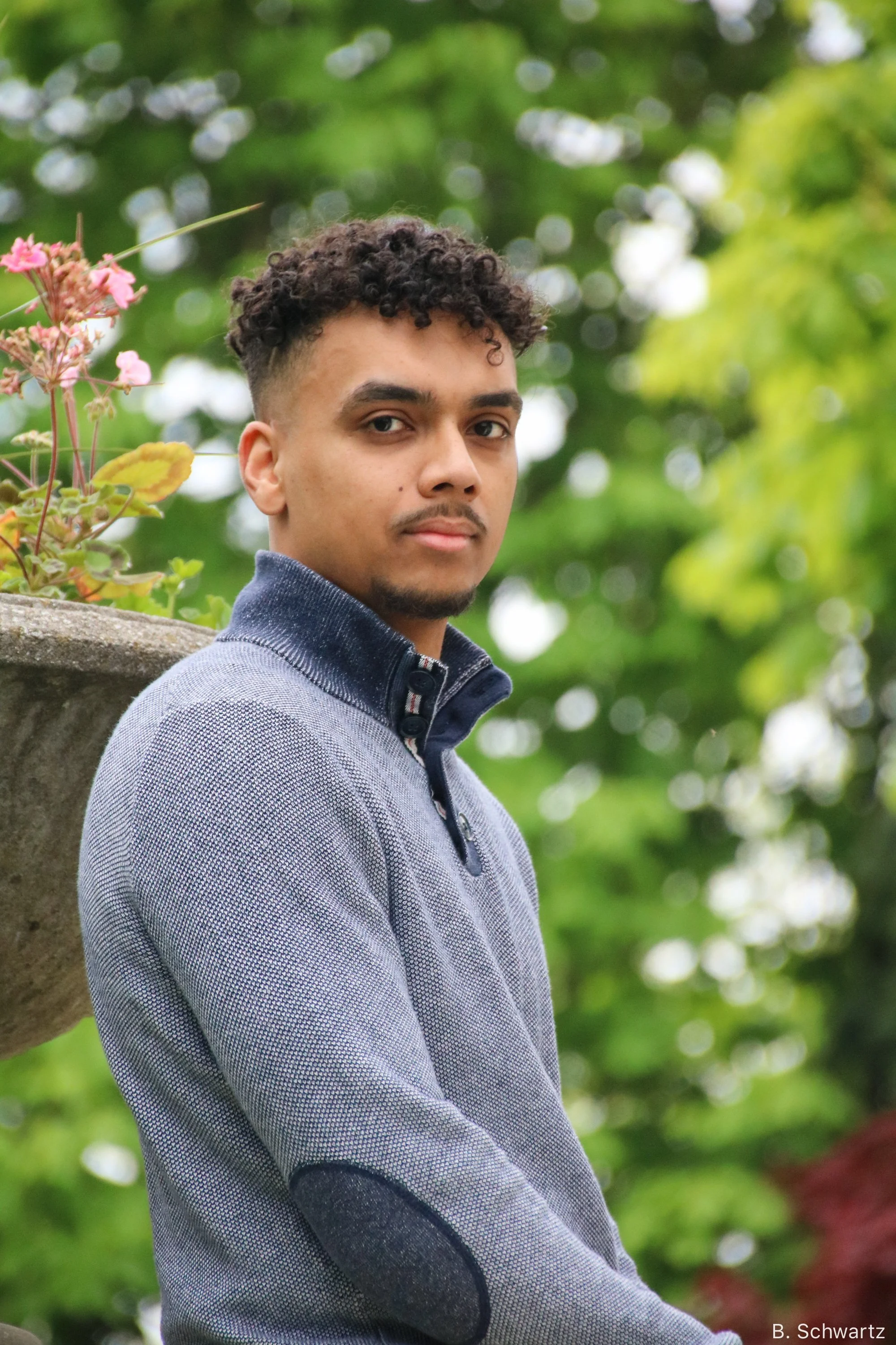A professional corporate headshot of a young man with curly hair and light brown skin standing outdoors next to a concrete planter with pink flowers, with lush green trees in the background.