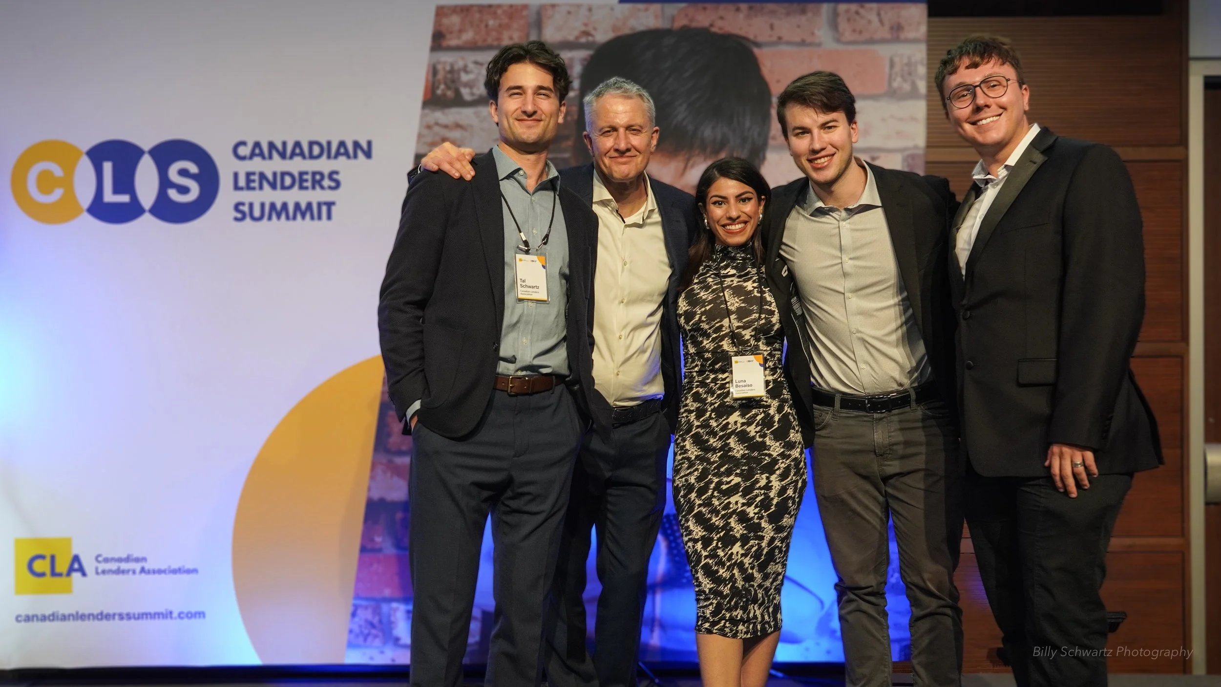 Group of five people smiling at the Canadian Lenders Summit, three men and one woman, standing in front of a backdrop with event logo and website.