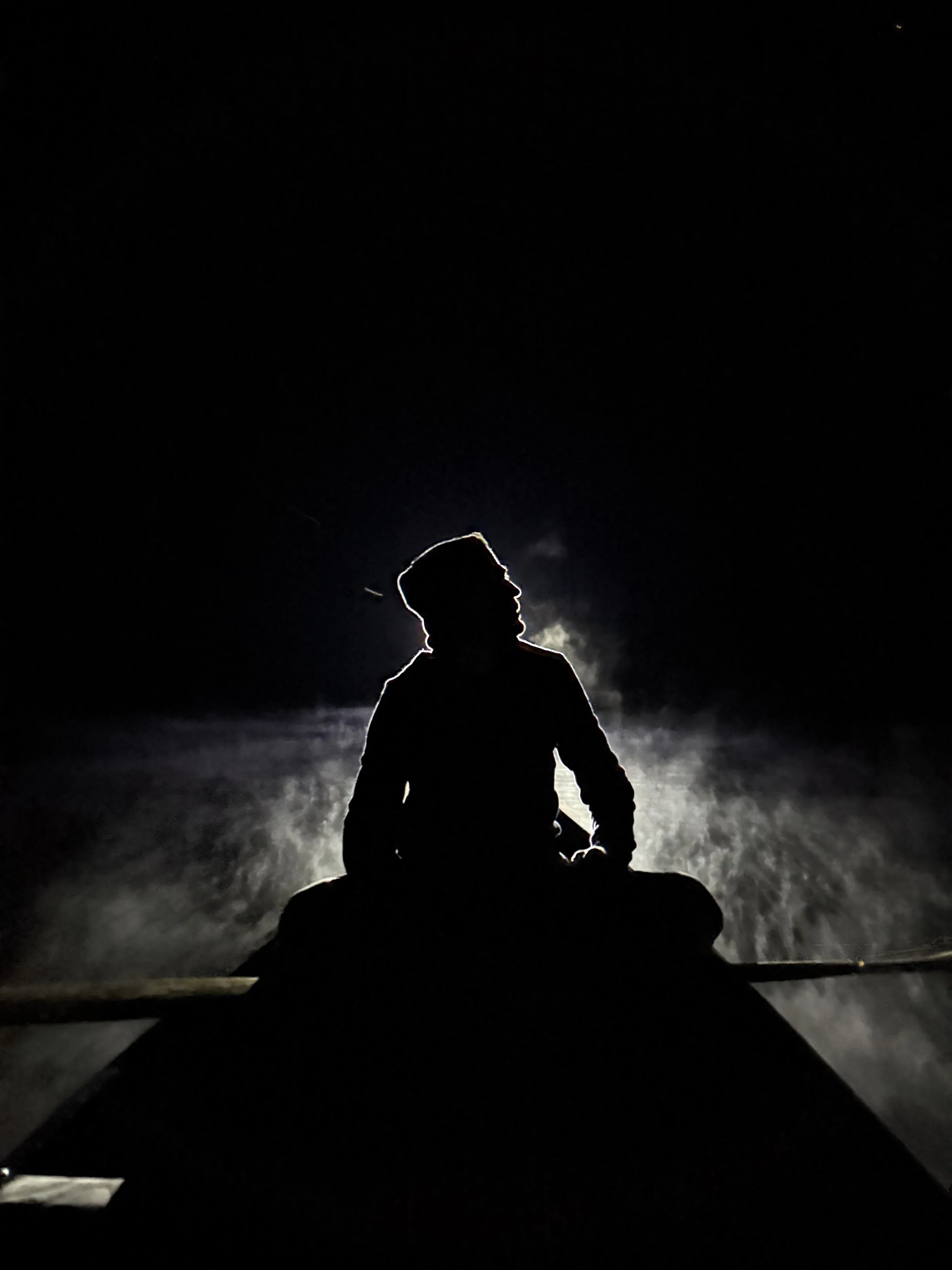 Silhouette of person sitting at the front of a boat at night with water spray behind them, illuminated from behind by a light source.