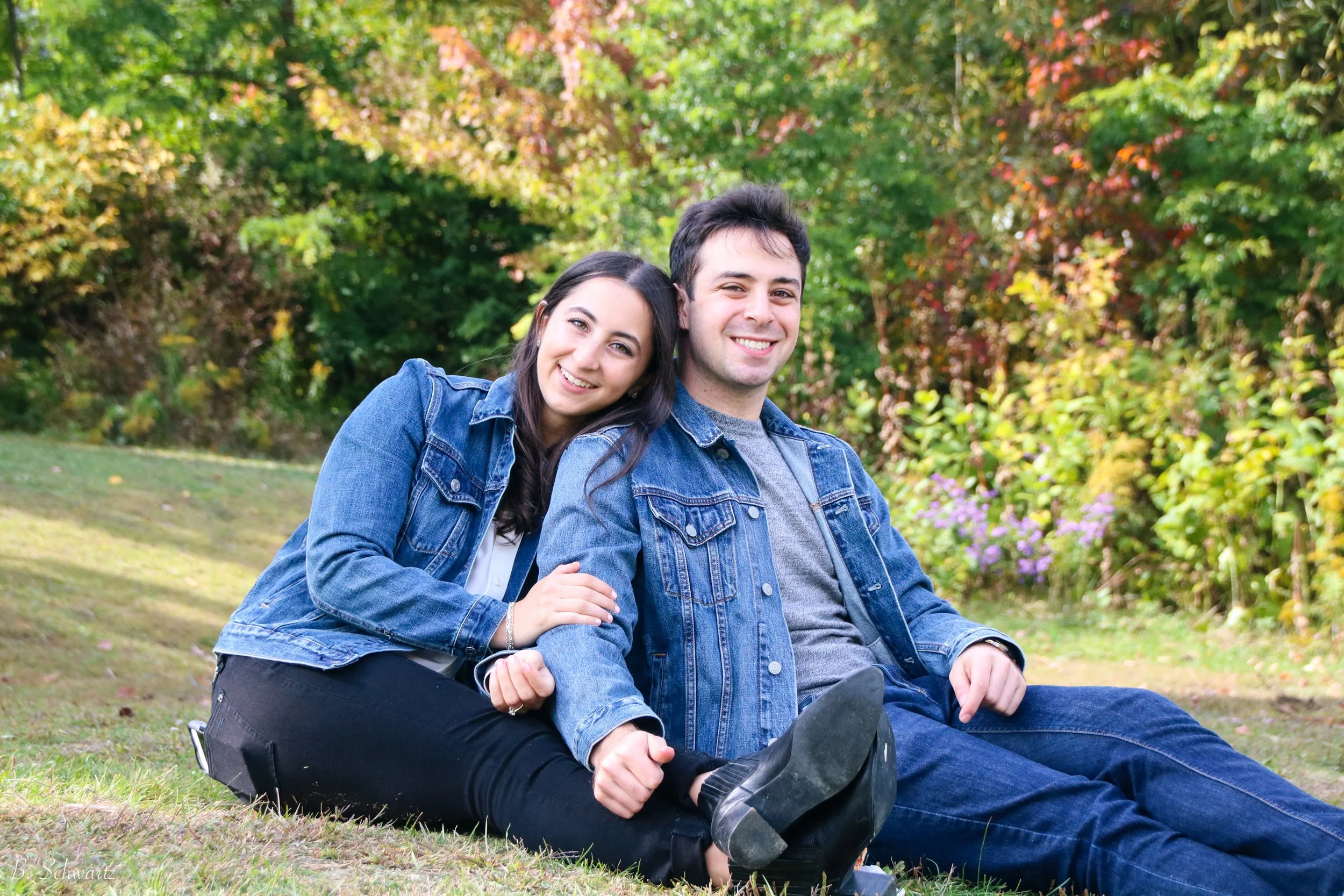 A young woman and a young man sitting on the grass in a park, smiling at the camera, with trees and colorful foliage in the background.
