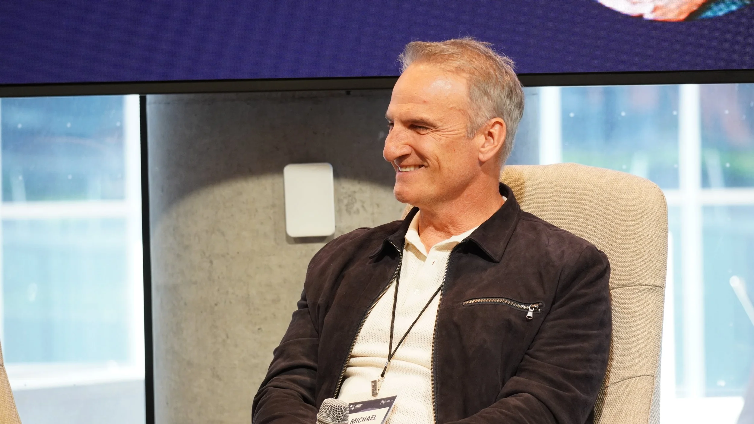 A middle-aged man with short gray hair smiling, sitting in a beige chair, wearing a black jacket and white shirt, with a name tag that reads 'Michael', holding a microphone, in a room with large windows in the background.