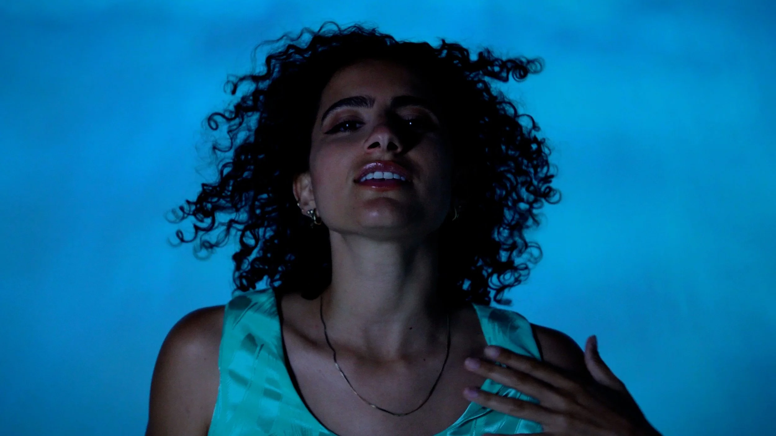 Woman with curly hair and earrings, wearing a teal top, with her hand on her chest, dark blue background.