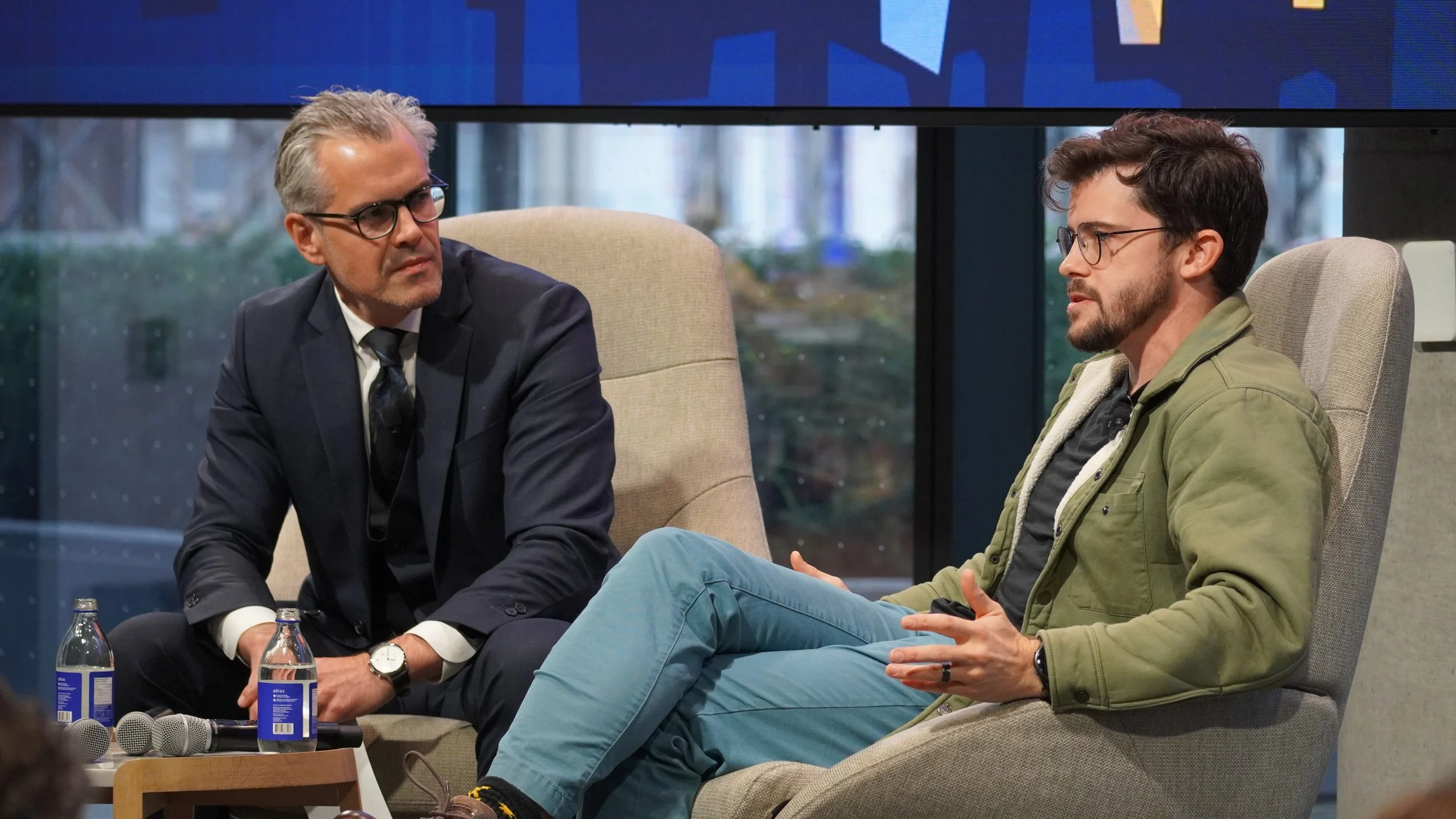 Two men sitting in chairs having a conversation, with microphones and water bottles on a small table nearby, set against a backdrop of glass windows with a cityscape.